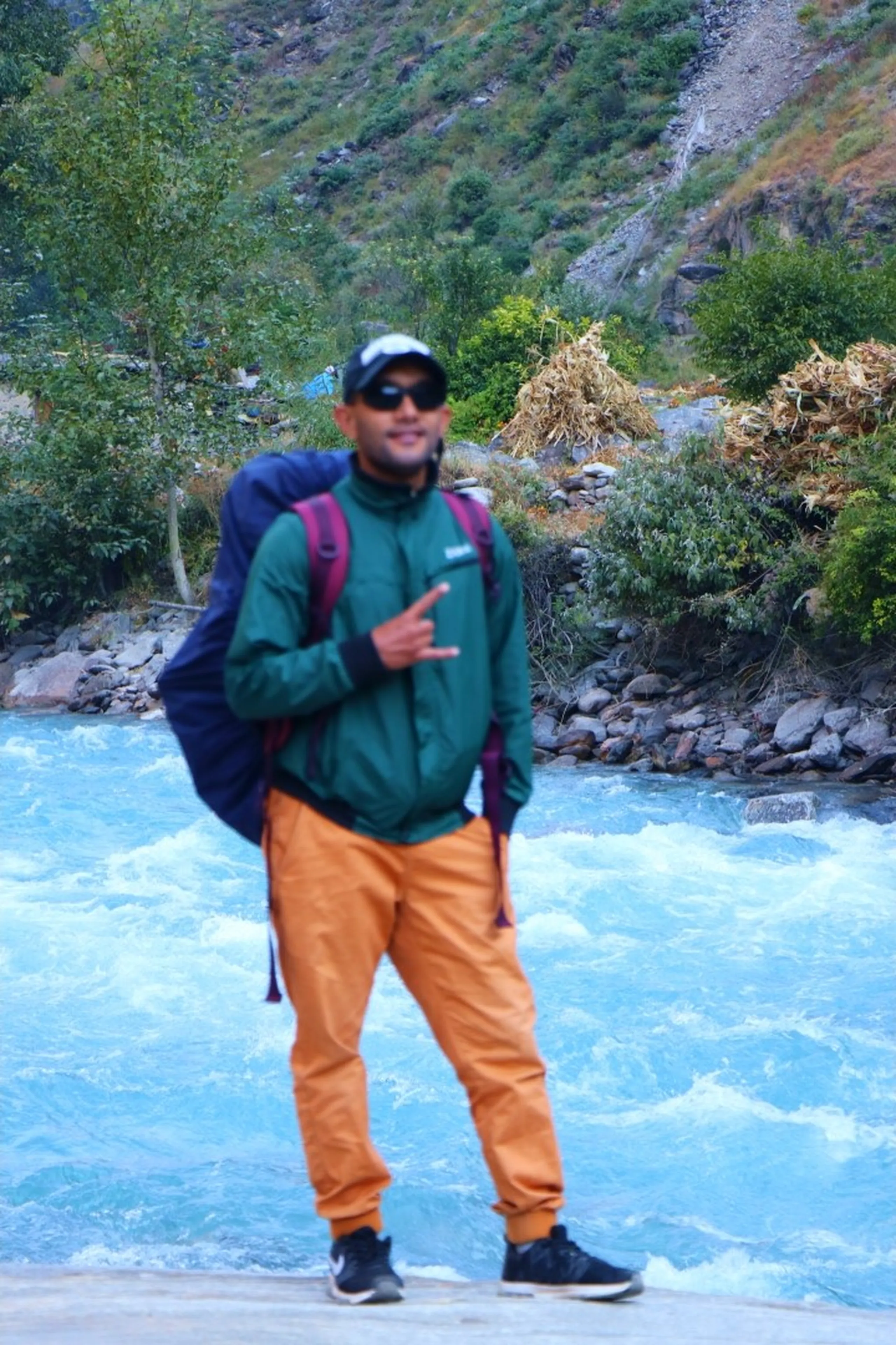 A slightly out-of-focus male trekker posing casually in front of a powerful rushing turquoise glacial mountain river, wearing a dark green zip-up fleece jacket, brown/tan jogger-style cargo pants, a white-trimmed black baseball cap and dark sunglasses with a backpack and dark jacket draped over one shoulder, giving a casual hand gesture toward the camera, lush green riverbanks with boulders and dense trees visible on the far side, Nepal river trekking, Shey Phoksundo trek river crossing, Himalayan glacial river adventure, Nepal trekking blog photo, mountain river portrait Nepal, flute player Nepal trek