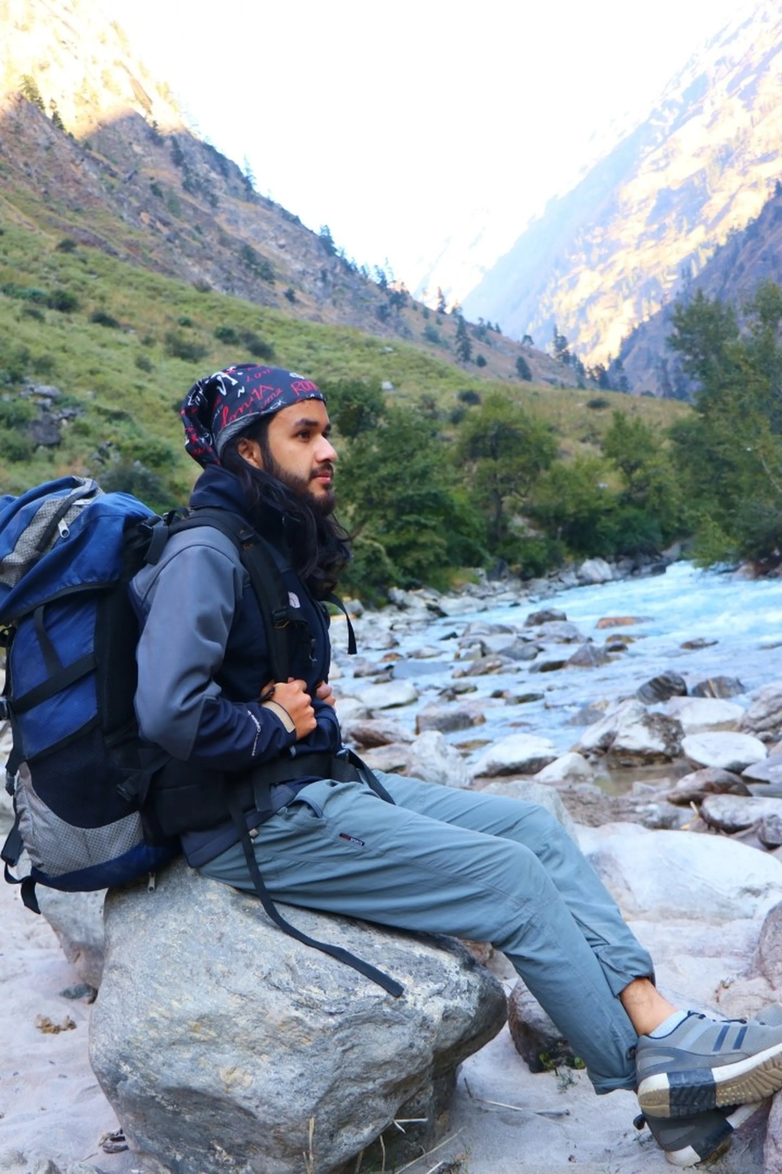 A bearded South Asian young male trekker sitting peacefully on a large white river boulder in profile view, wearing a patterned dark bandana on his head, a grey-blue soft shell trekking jacket layered under a North Face vest, light grey trekking pants and grey Adidas trail shoes, carrying a large blue and grey hiking backpack, gazing contemplatively upstream along a stunning turquoise glacial river flowing through a narrow Himalayan gorge flanked by snow-dusted rocky peaks under a bright sky, solitary trekker Nepal mountains, Shey Phoksundo river rest stop, Himalayan trek meditation moment, Nepal wilderness photography, trekking aesthetic Nepal