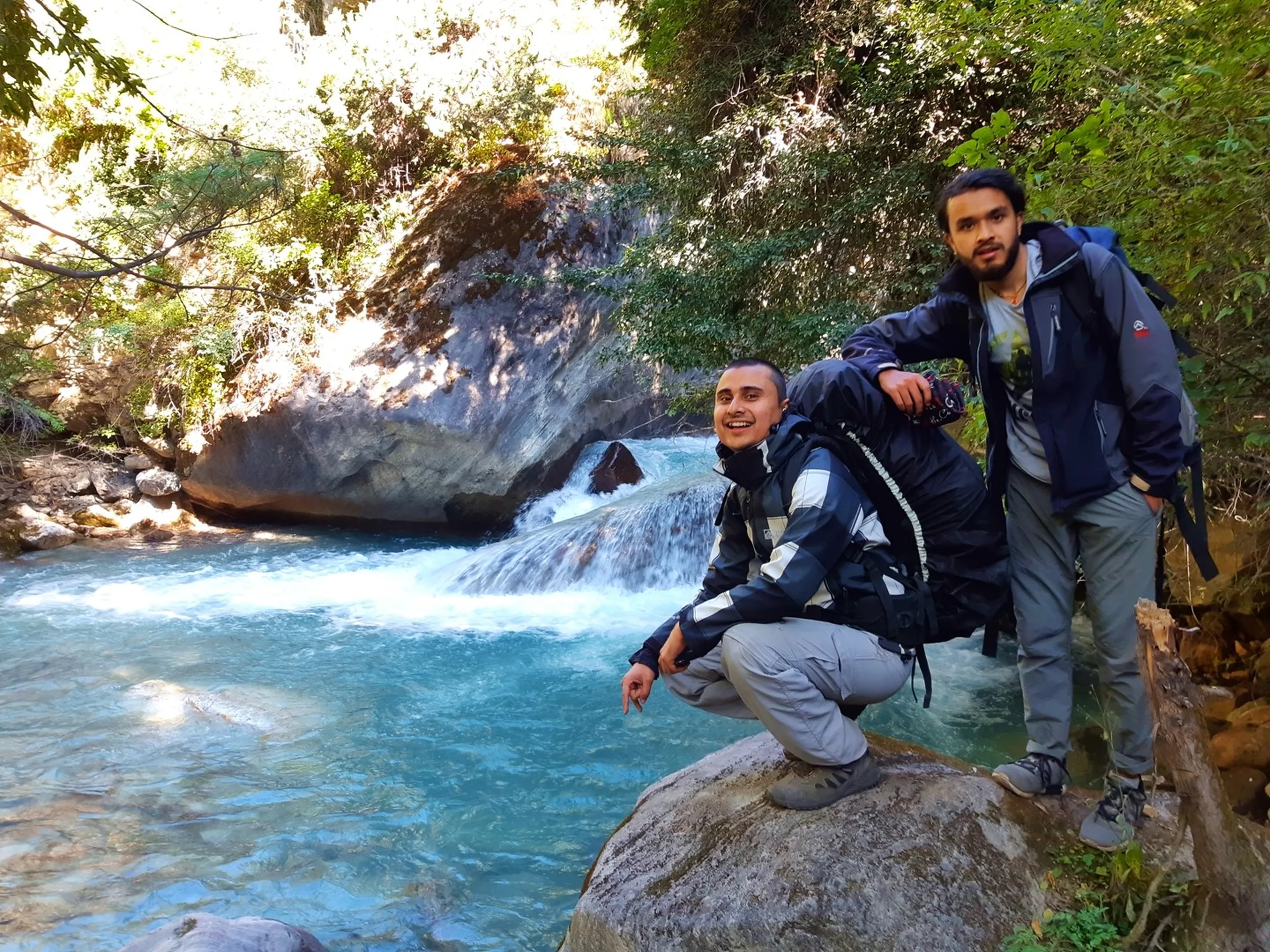 Two South Asian male trekkers posing beside a serene crystal-clear turquoise mountain stream waterfall in a forested gorge - the shorter one with a buzz cut crouching on a smooth boulder in a dark grey and silver windbreaker jacket and light grey trekking trousers with a large backpack, smiling broadly at the camera, while the taller one stands confidently behind him in a dark hiking jacket and grey trousers with his own backpack, lush green overhanging foliage framing the scene, smooth water-worn white boulders and bright blue-green pool visible, Nepal hidden waterfall trek, Shey Phoksundo trekking scenery, Himalayan mountain stream photography, Nepal nature trekking, backpackers waterfall Nepal