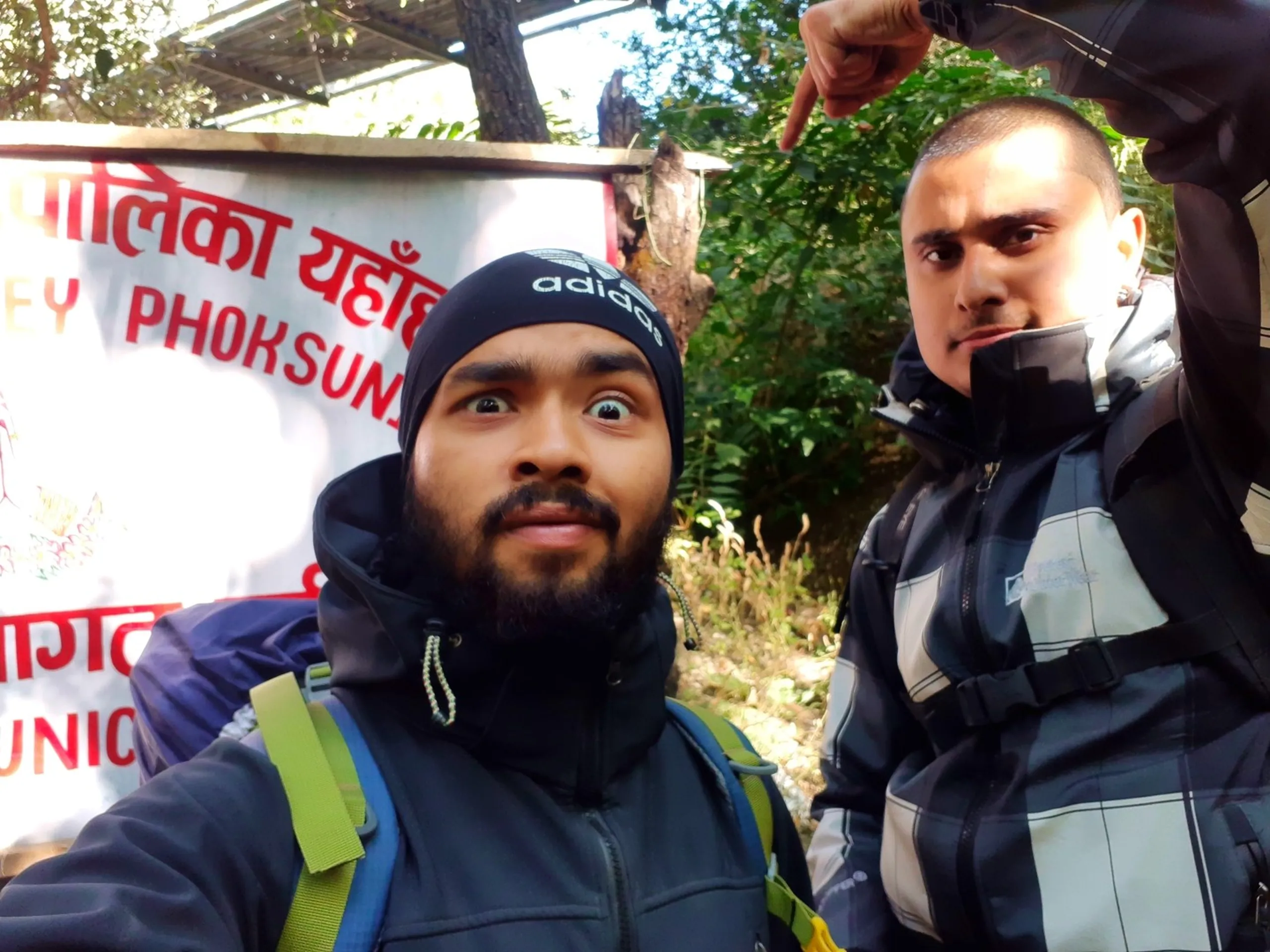 A close-up selfie of two young South Asian male trekkers in a forest area beside a Nepali government or municipality sign board with red Devanagari text and partial English text reading "PHOKSUN..." (Phoksundo), one in the foreground wearing a black Adidas beanie hat and dark puffer jacket with a green-strapped blue backpack making a wide surprised bug-eyed expression directly at camera, the other partially visible behind in a black and grey riding or trekking jacket gesturing with a finger pointing downward toward his companion, playful and humorous portrait, Shey Phoksundo sign board Nepal trek, funny trekking selfie Himalaya, Nepal national park entrance, Phoksundo trek checkpoint, batch mates Nepal adventure
