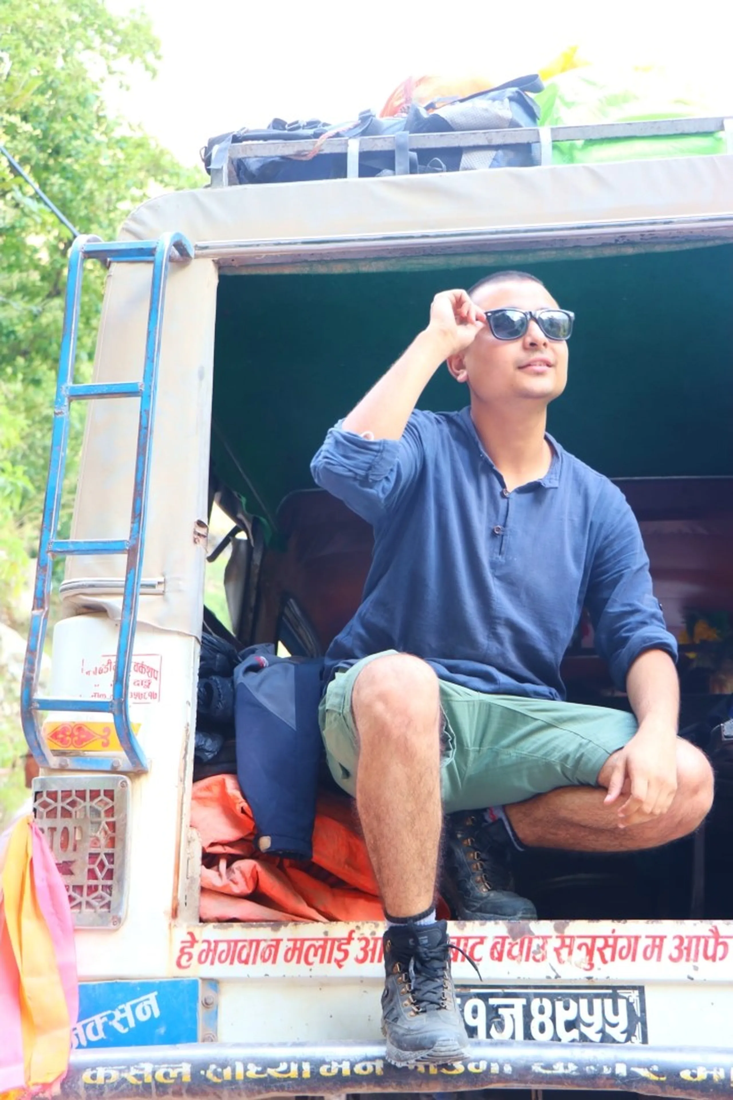 A young South Asian male trekker sitting coolly at the open rear door of a decorated Nepali Bolero jeep, wearing dark navy blue long-sleeve shirt, olive green cargo shorts, black lace-up hiking boots and black sunglasses, one hand adjusting his shades while gazing sideways with a relaxed expression, gear and bags visible on the jeep's rooftop rack above him, lush green trees blurring in the background, Nepali Devanagari script visible on the vehicle including a license plate reading "9J 4Y55", adventure travel Nepal, Himalayan road trip, jeep travel Nepal mountains, Shey Phoksundo trek journey