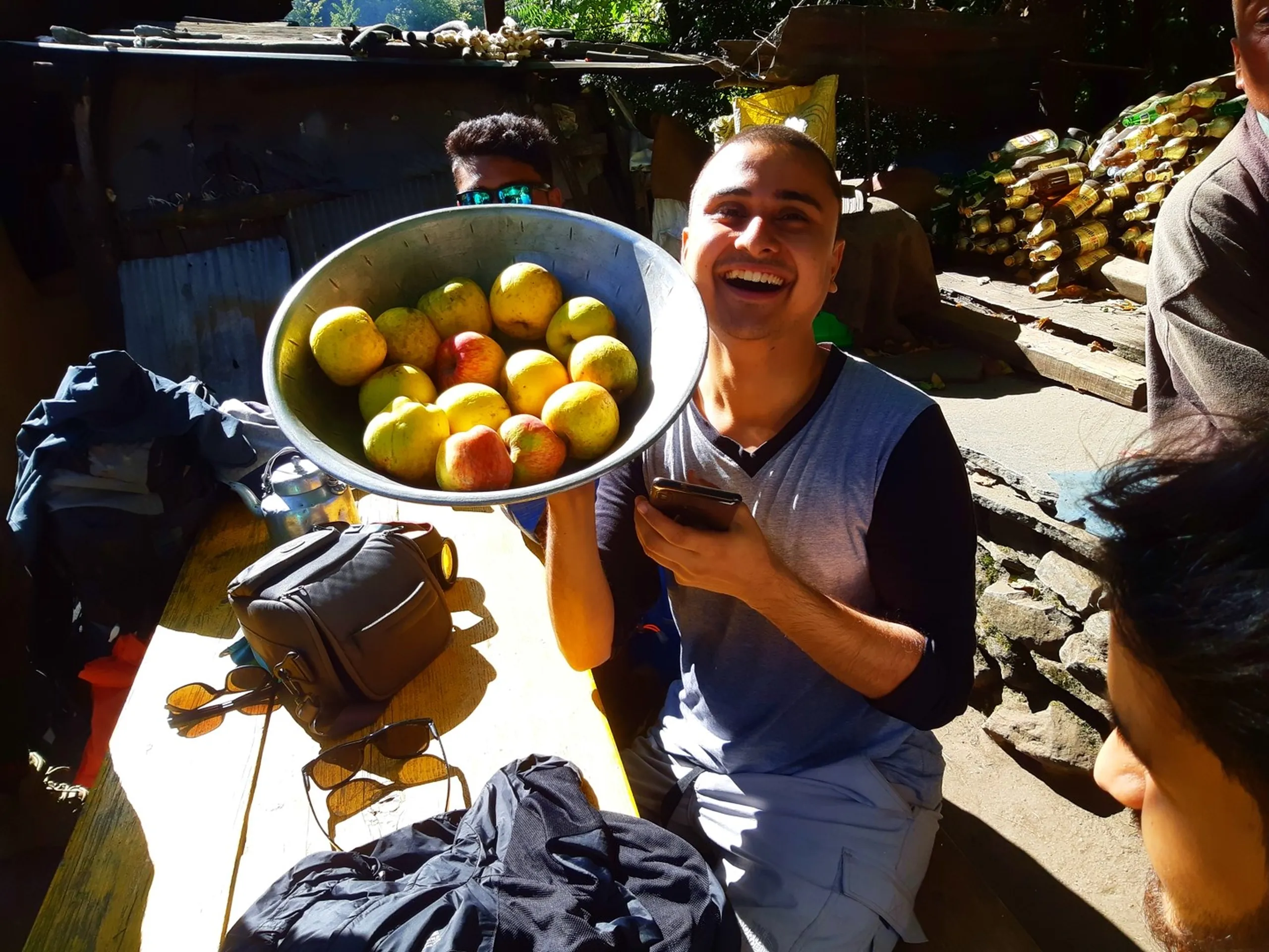 A young South Asian male trekker with a shaved buzz cut and a wide infectious laugh holding up a large round aluminum plate or tray piled high with fresh locally grown yellow-green apples and a few red apples, wearing a grey and black short-sleeve shirt and light grey trekking trousers while seated outdoors at a rustic Nepali village market stall, another trekker with teal sunglasses partially visible behind him, rows of plastic bottles stacked in the background, sunlight casting dramatic shadows, fresh local fruit Nepal trek, Shey Phoksundo apple season Nepal, Himalayan village market trekking, Nepal fresh apples mountains, authentic Nepal trekking experience, local produce Nepal village