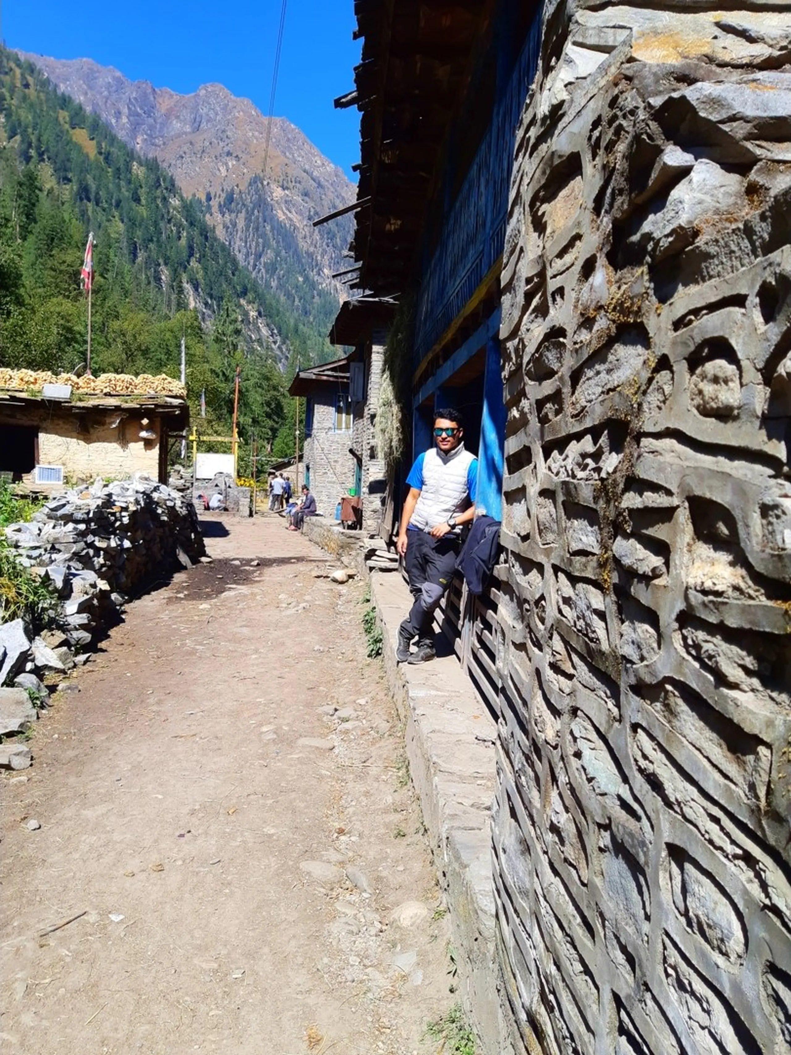 A young South Asian male trekker leaning casually against the intricately stacked grey stone wall of a traditional Nepali mountain village building, wearing a white quilted sleeveless vest over a bright blue t-shirt, dark trekking trousers and black hiking shoes with teal mirrored sunglasses, positioned along a narrow dusty trekking trail flanked by ancient stone-walled structures with weathered wooden balconies painted blue, corn cobs drying on a rooftop visible to the left, a Nepali flag fluttering in the background, tall green pine-forested Himalayan peaks rising under a clear deep blue sky, traditional Nepali stone village architecture Shey Phoksundo trek route, remote Himalayan village Nepal, college friend trekking Nepal, Dolpa district Nepal travel photography, authentic mountain village life Nepal