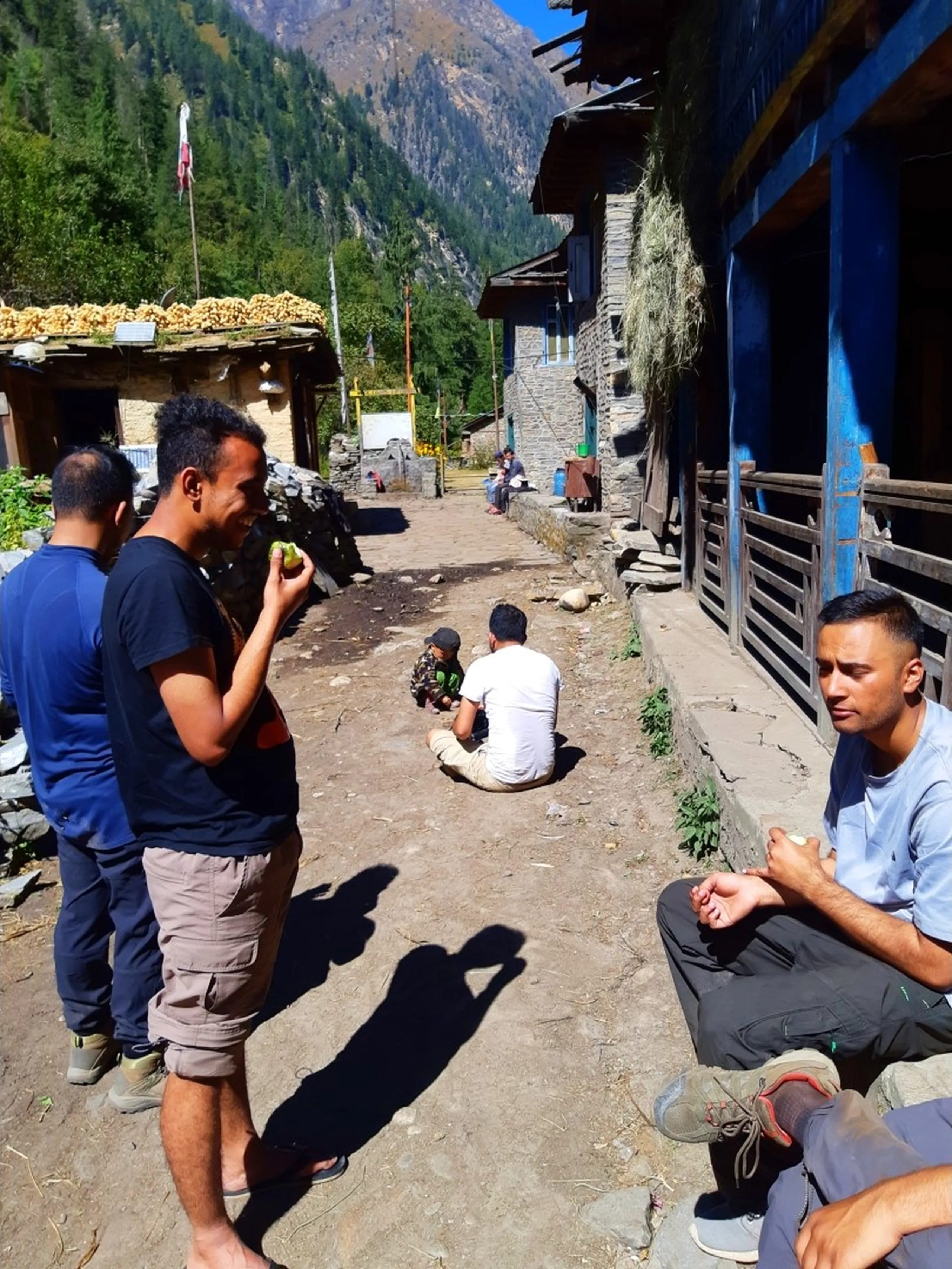 A candid wide-angle scene of a diverse trekking group taking a rest break in the dusty central lane of a remote traditional Nepali stone village at Chepka, surrounded by rustic blue-painted wooden-balconied buildings with hay bundles hanging from the eaves and corn drying on rooftops - one bearded young man in a black t-shirt and khaki shorts stands eating a green fruit, another in a navy blue jacket partially visible to the left, a third in a white t-shirt sits cross-legged on the ground with a local child nearby, while a fourth in a light grey t-shirt sits on a stone ledge in the foreground, prayer flags and a Nepali flag visible in the distance, steep green forested Himalayan peak towering under a clear blue sky, trekking rest stop Nepal remote village, Chepka village Shey Phoksundo trek, batch mates Nepal mountain break, authentic Himalayan village community, Dolpa trekking Nepal blog