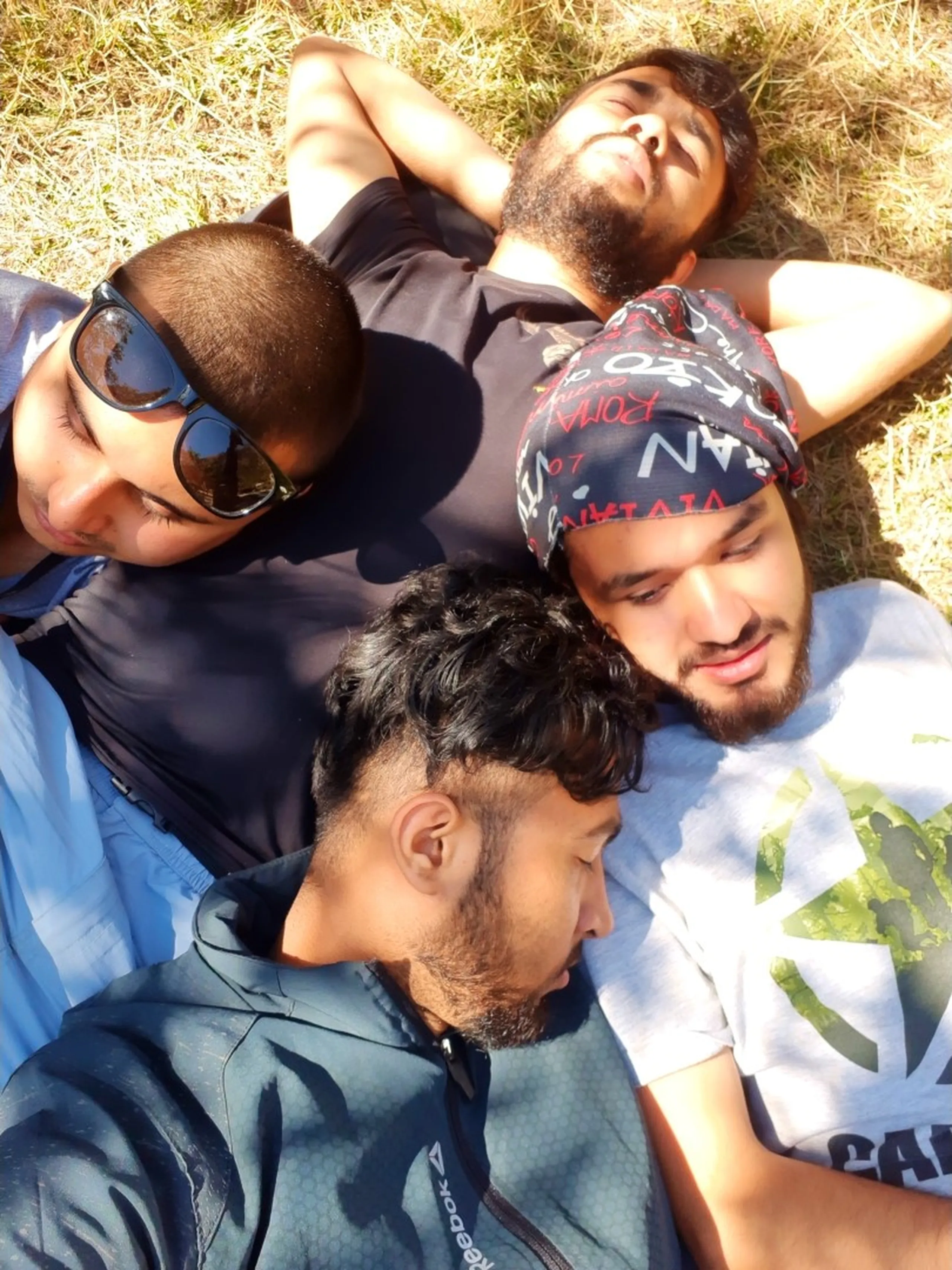 An overhead top-down close-up portrait of four exhausted but content young South Asian male trekkers lying head-to-head on dry golden mountain grass resting after a long tiring day on the trail, arranged in a circle formation - one with a buzz cut and dark sunglasses reflecting the sky on the left, one in a black t-shirt with eyes closed and arms behind his head at the top, one with a patterned black and red bandana and a light beard wearing a white Reebok graphic t-shirt on the right, and one in a dark grey Reebok zip jacket in the foreground with curly dark hair looking downward, all showing signs of joyful post-hike exhaustion, sunlight casting strong shadows on dry alpine grassland, Shey Phoksundo trek rest Nepal, batch mates mountain nap Nepal, exhausted trekkers Nepal Himalaya, college friends trek bonding Nepal, Dolpa trekking friends circle