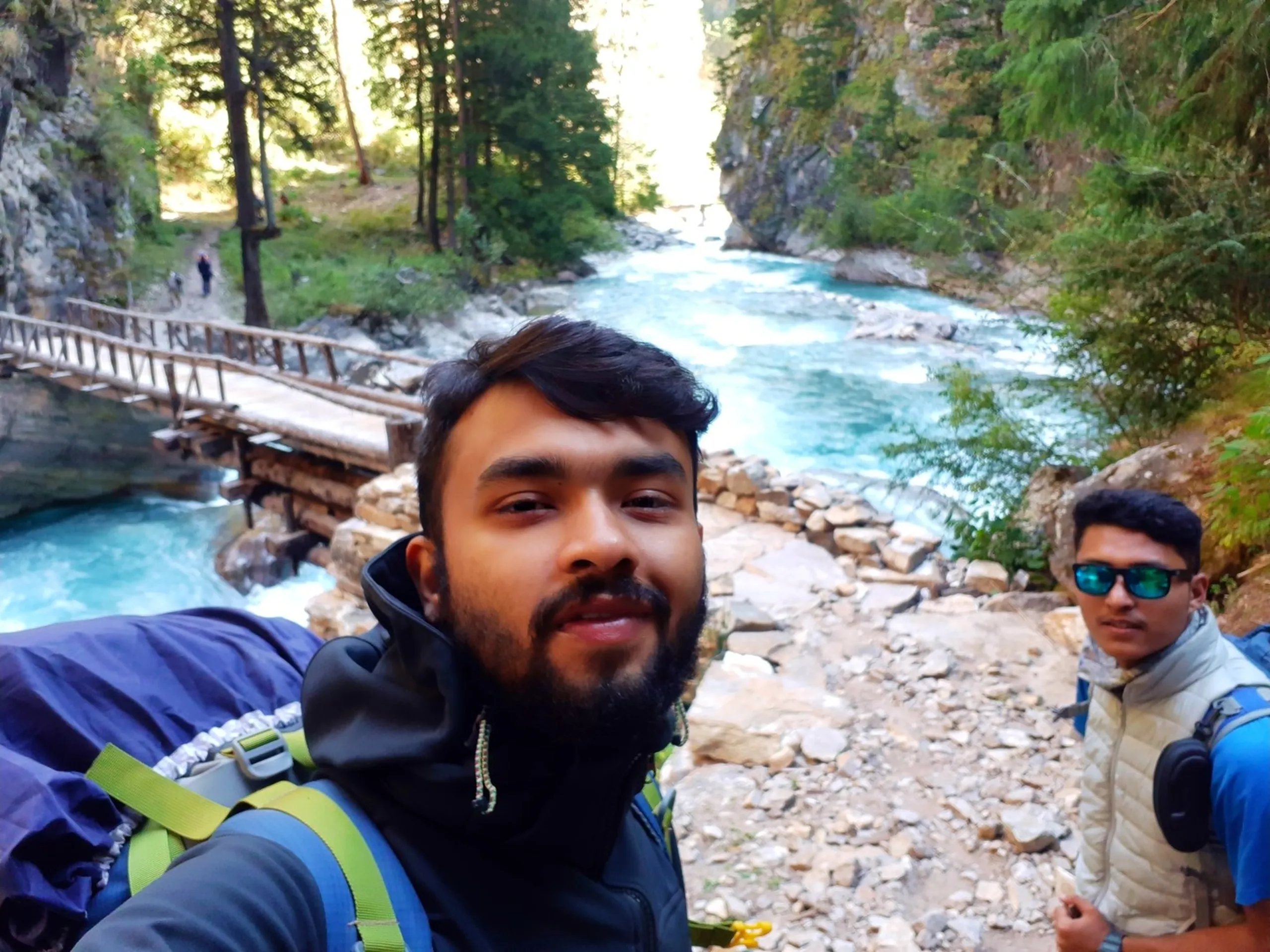 A close-up trekking selfie taken by a bearded South Asian male trekker wearing a dark hoodie jacket with a large green-strapped blue backpack, smiling warmly directly at the camera, with his batch mate in a white sleeveless vest and blue t-shirt with teal sunglasses partially visible behind him to the right - both standing on a rocky trail beside a dramatic turquoise glacial river gorge lined with towering dark pine trees, a rustic wooden plank footbridge crossing the rushing river visible in the middle distance with tiny silhouettes of trekkers on it, steep rocky canyon walls rising on either side under an overcast forest canopy, Nepal wooden bridge trekking selfie, Shey Phoksundo trek trail wooden bridge, glacial river gorge Nepal Himalaya, college batch mates trekking Nepal, Dolpa trek adventure photography