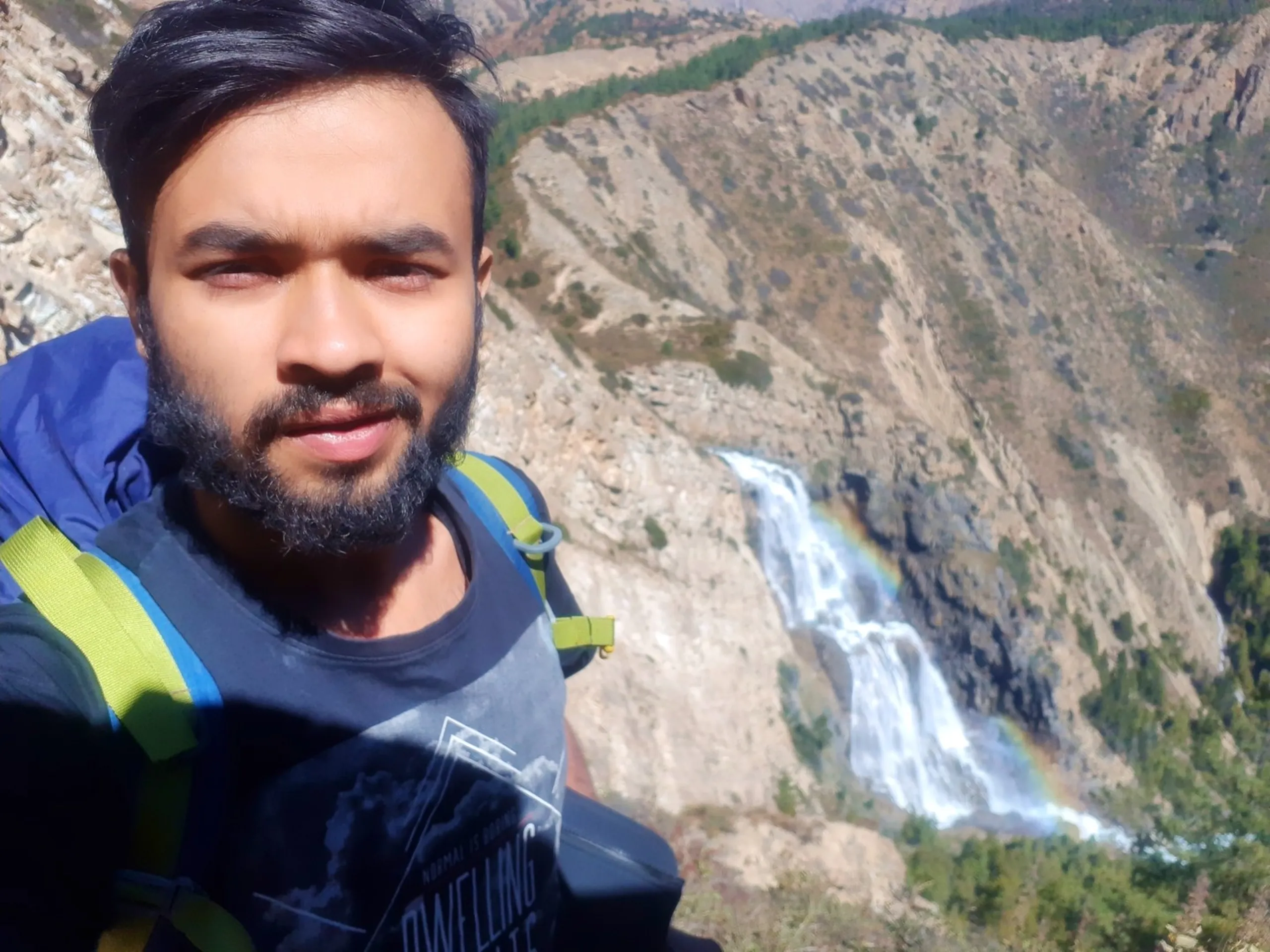 A close-up trekking selfie taken by a bearded South Asian male trekker in a dark graphic t-shirt with a large blue backpack with lime green straps visible on his shoulder, wearing a subtly confident smirk, with a breathtaking dramatic Himalayan waterfall cascading in multiple tiers down a sheer bare rocky cliff face directly behind him in the background, a faint natural rainbow visible in the mist at the base of the waterfall, sparse green shrubs dotting the rocky slopes, Shey Phoksundo waterfall Nepal selfie, Himalayan waterfall trek Nepal, rainbow waterfall Nepal Dolpa, trekker selfie waterfall Nepal, Shey Phoksundo National Park trek photography, most beautiful waterfall Nepal Himalaya
