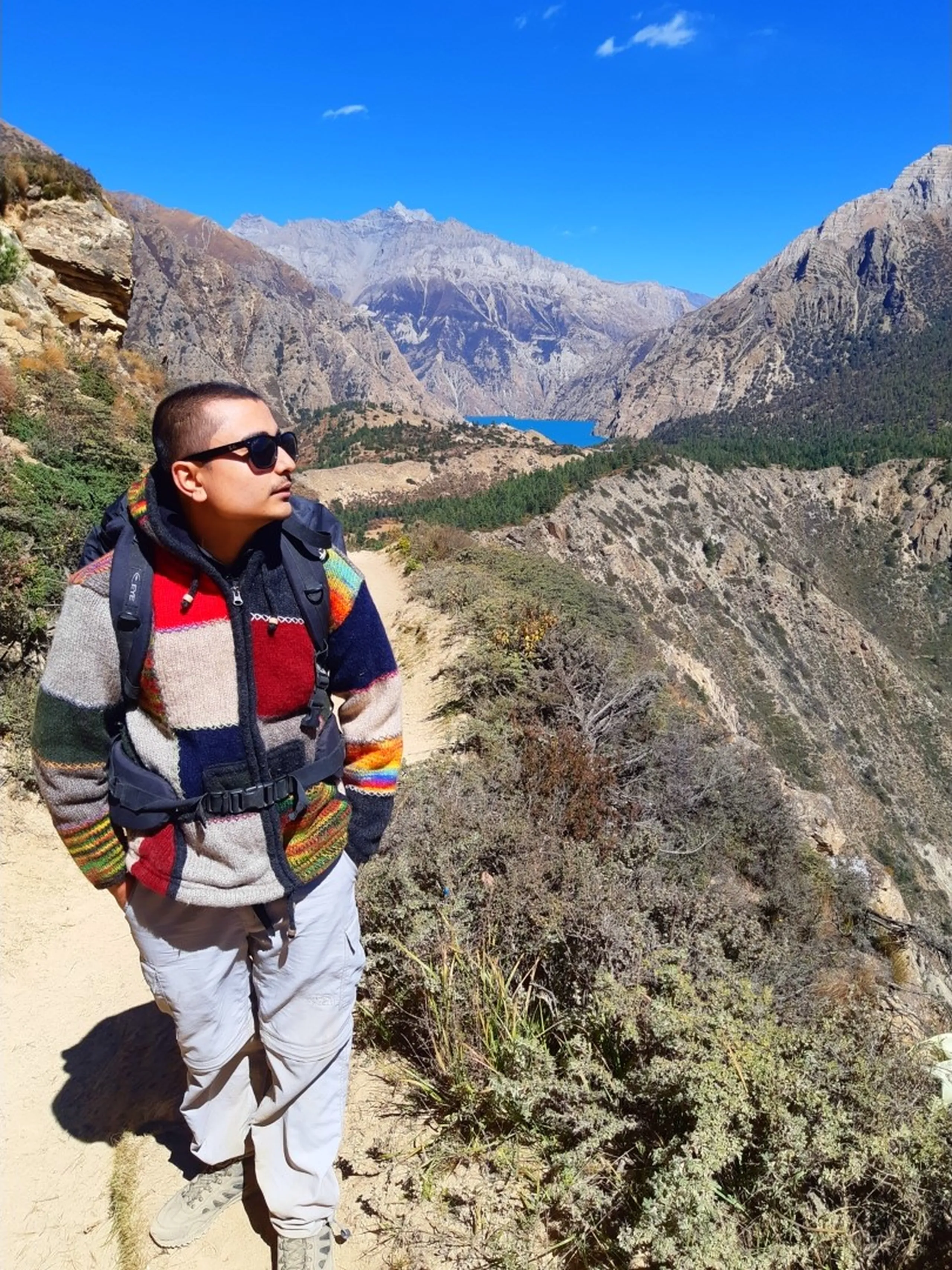 A stylish male trekker with a buzz cut and dark sunglasses standing on a narrow exposed dusty mountain trail, wearing a vibrant multicolored traditional Nepali wool knit sweater with bold horizontal stripes of red, navy, blue, grey and yellow with a dark backpack vest harness over it and light grey North Face trekking trousers and hiking shoes, hands tucked casually in pockets, gazing sideways away from camera with a thoughtful faraway expression, an absolutely jaw-dropping panoramic view behind him - a vivid turquoise Shey Phoksundo Lake nestled between massive barren grey Himalayan ridges under a perfectly clear deep blue sky, Shey Phoksundo Lake Nepal view, Dolpa Nepal trekking panorama, turquoise alpine lake Nepal Himalaya, traditional Nepali wool sweater trekking, Shey Phoksundo National Park vista, best views Nepal trek