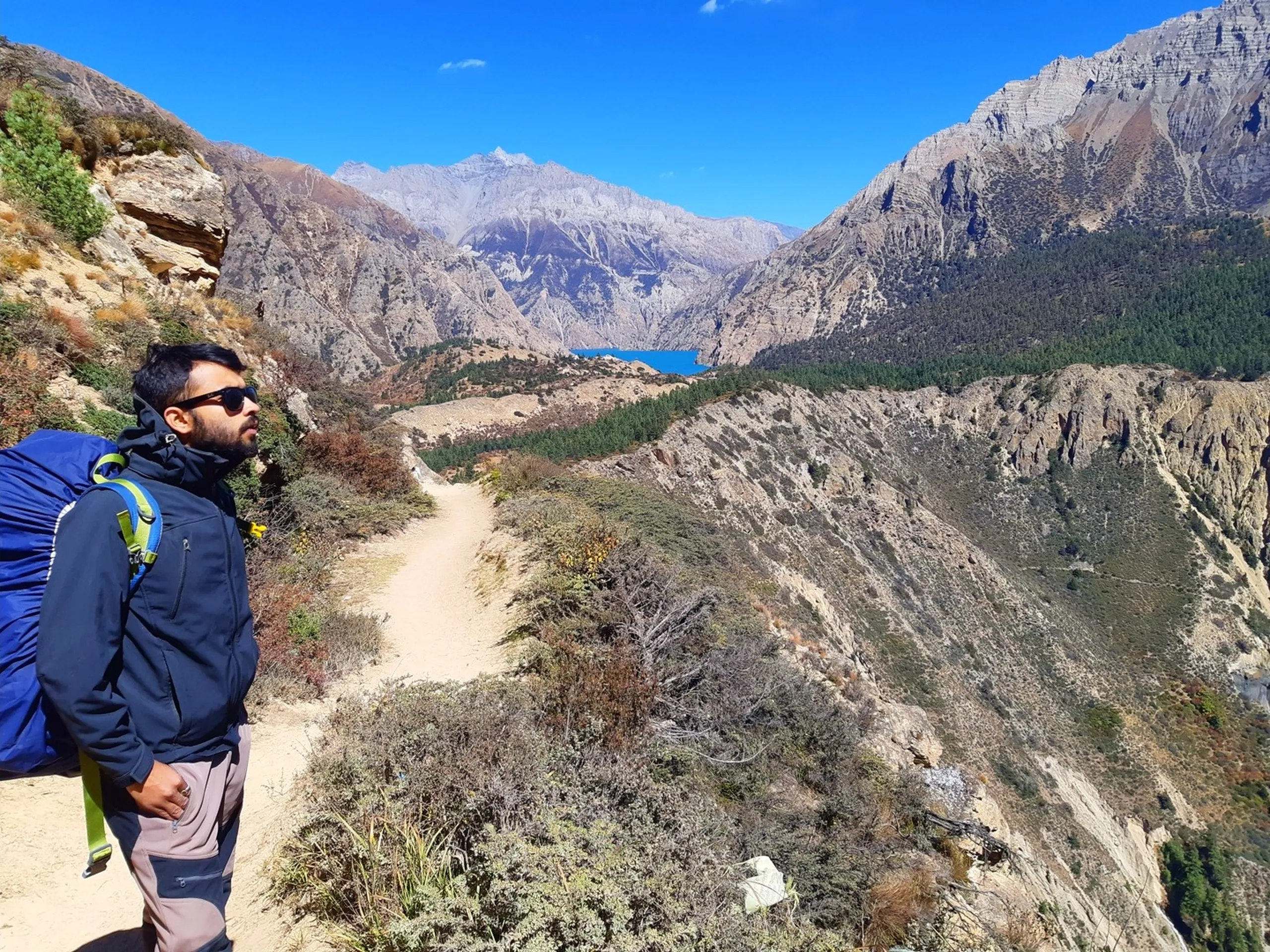 A contemplative bearded South Asian male trekker with long dark hair tied back standing in profile view on a narrow cliff-edge mountain trail, wearing a dark blue zip-up softshell jacket and light grey trekking trousers with a large blue and grey backpack, one hand grasping a yellow strap, gazing out thoughtfully over a sweeping arid Himalayan valley - a winding pale sandy trail descending through scrubby brown bushes toward the vivid turquoise expanse of Shey Phoksundo Lake far below, framed between towering naked barren grey Himalayan peaks under a rich blue sky, a crashed or discarded piece of metalwork barely visible on the precipitous cliff edge below, Shey Phoksundo Lake approach trail Nepal, Dolpa trek viewpoint Nepal, male trekker Himalayan cliff trail, Shey Phoksundo National Park trekking Nepal, best treks Nepal high altitude lake