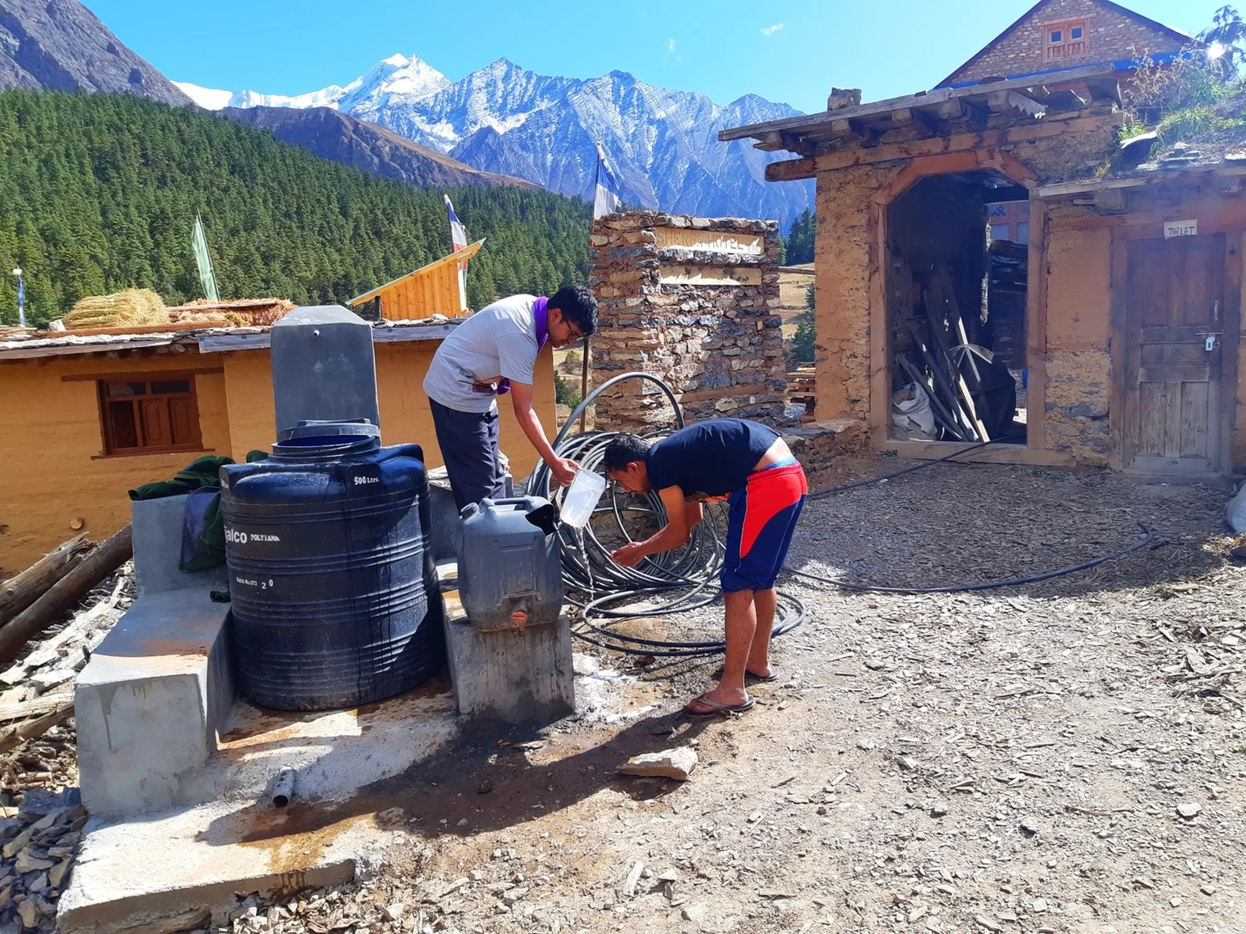 Two young South Asian male trekkers washing up at a simple outdoor water tank station in a remote Nepali mountain village, surrounded by the stark contrast of mundane plastic black water storage tanks labeled "Italco Polytank 500 Litres" against a breathtaking backdrop of towering snow-capped Himalayan peaks under a clear blue sky - one taller trekker in a white t-shirt and glasses standing and carefully pouring water from a clear plastic container, while the other in a black t-shirt and red board shorts bends over washing his hands or face at the spigot, coiled black irrigation hoses scattered around, an old stone and mud building labeled "Toilet" visible to the right, Buddhist prayer flags fluttering in the breeze, remote Nepal village water source trek, Shey Phoksundo trekking camp hygiene, batch mates morning wash Nepal trek, Himalayan village life Nepal, snow peak backdrop Nepal trek camp