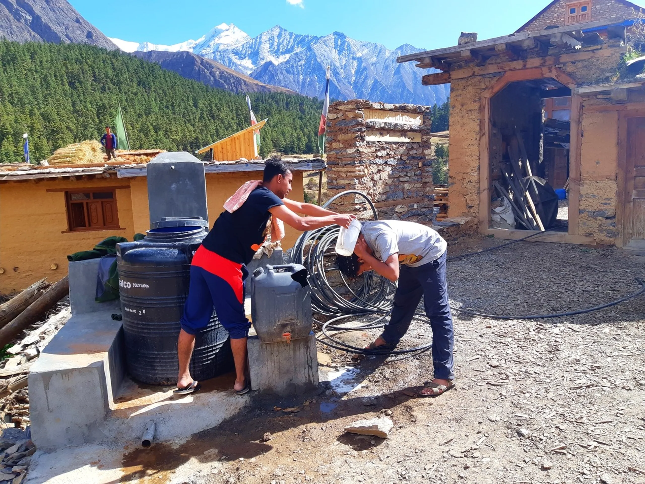 The same two young male trekkers at the same outdoor water station now reversed in roles - the one in the black t-shirt and red board shorts now standing and dramatically pouring a full container of water directly over the bent-forward head of his companion in the white t-shirt who washes his hair while bracing against the stone pillar, a towel draped over the standing trekker's shoulder, both barefoot in flip flops, the scene playfully capturing the camaraderie of washing hair in a remote high-altitude Himalayan village with snow-dusted peaks still dominating the background, a person visible on a rooftop in the far left, Nepal trek outdoor hair wash, high altitude cold wash Himalaya, trekking friends camp life Nepal, Shey Phoksundo trek base camp activities, remote Nepal mountain camp hygiene
