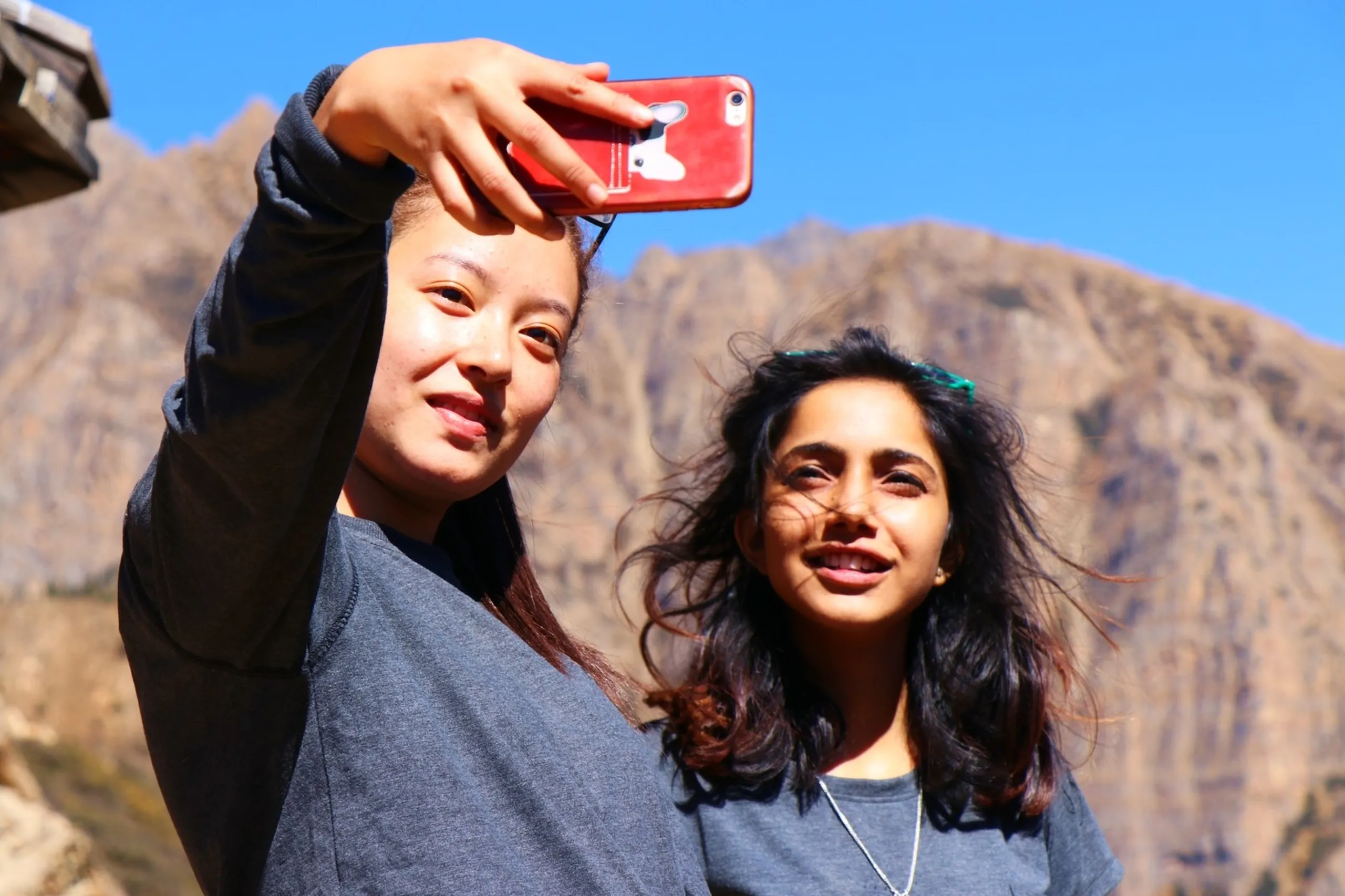 Two young South Asian women taking a cheerful selfie together against a dramatic high-altitude Himalayan mountain backdrop under a crisp clear blue sky - the one on the left holding a red iPhone case up at arm's length to capture the shot, wearing a dark grey-black long-sleeve top with hair neatly pulled back, while the one on the right smiles warmly with wind-tousled curly dark hair, wearing a grey t-shirt with a delicate silver pendant necklace and teal hair clips, jagged bare brown Himalayan rocky ridgeline framing the background, female trekkers selfie Nepal Himalaya, women trekking Shey Phoksundo Nepal, college batch mates female trek Nepal, girl friends mountain adventure Nepal, Dolpa trek women empowerment, Nepal high altitude trek selfie