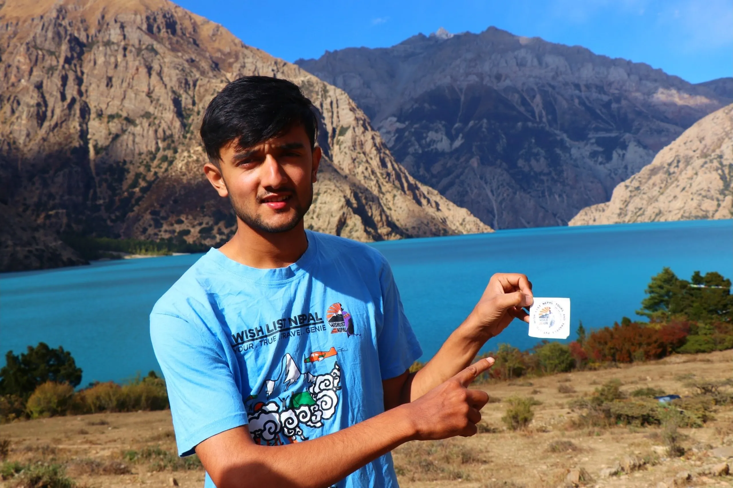 A young South Asian male trekker with a neatly trimmed short beard and dark hair smiling proudly while holding up and pointing to a small square sticker or badge of his travel company logo between his fingers, wearing a light blue graphic t-shirt printed with "Wish List Nepal - Your True Travel Genie" text and a colorful adventure-themed illustration, standing before the most iconic scene of the entire trek - the vast luminous turquoise Shey Phoksundo Lake stretching across the background between towering barren rocky Himalayan cliffs under a brilliant blue sky, trekking company Nepal branding, Wish List Nepal travel agency Shey Phoksundo, Shey Phoksundo Lake Nepal destination reached, trekking milestone Nepal Dolpa, Nepal travel agency trek Himalaya, Shey Phoksundo National Park arrival