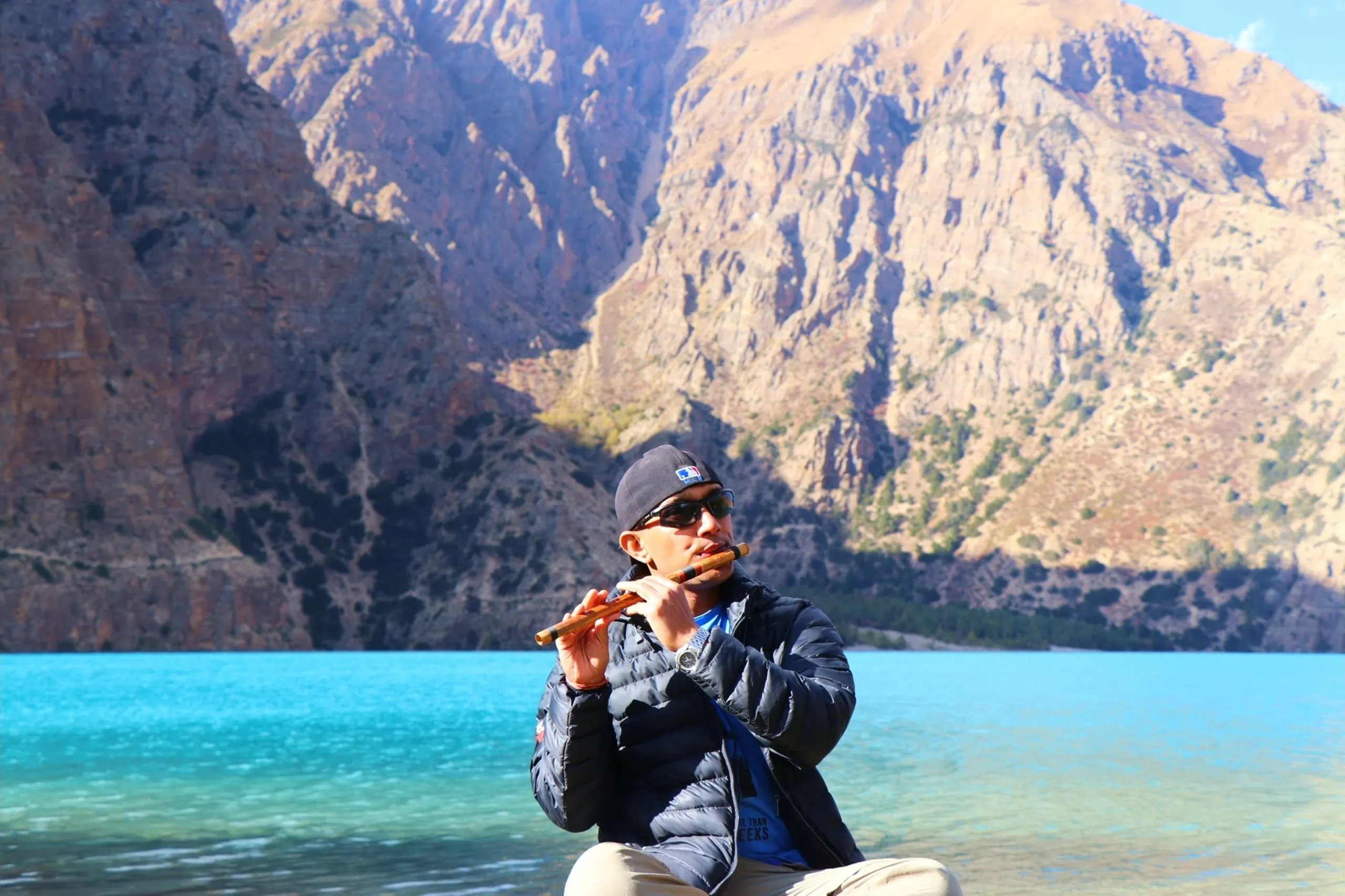 A serene and deeply atmospheric portrait of a male musician and trekker sitting on a low rock at the edge of the impossibly vivid turquoise Shey Phoksundo Lake, playing a traditional Nepali bamboo bansuri flute with both hands raised and eyes focused, wearing a black MLB-logo beanie hat, dark sunglasses, a black puffer down jacket over a blue t-shirt and beige trekking trousers, the massive dramatic barren rocky vertical cliff walls of the Himalayan canyon rising directly behind the still lake waters, the entire scene radiating peace and cultural harmony between music, man and mountain, flute player Shey Phoksundo Lake Nepal, bansuri flute Himalayan lake, Nepal music mountains, Dolpa lake Nepal cultural, traditional Nepali flute music trekking, Shey Phoksundo Lake serenity Nepal