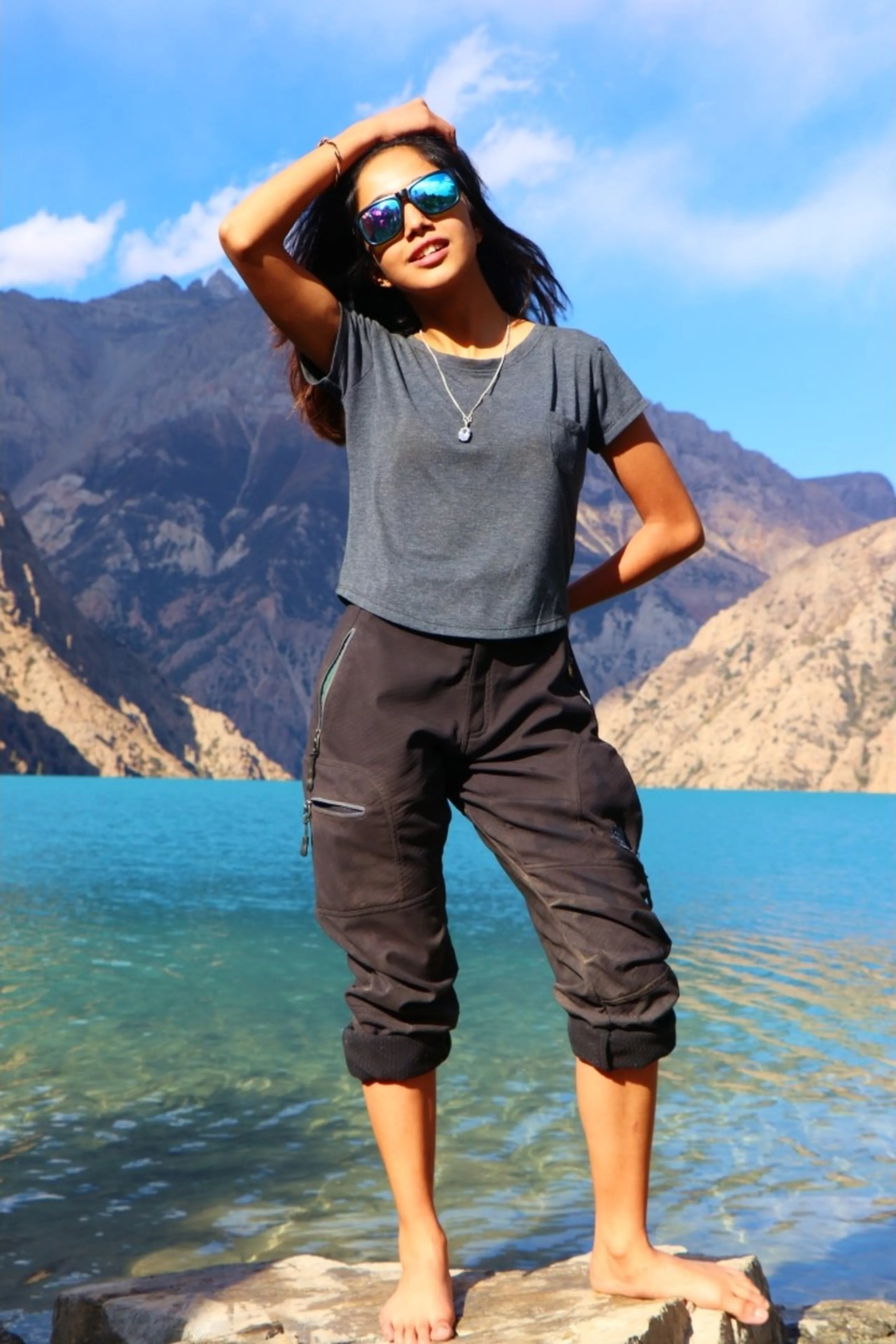 A striking and confident full-body portrait of a young South Asian woman standing barefoot on flat stones at the very edge of the crystal-clear shallows of Shey Phoksundo Lake, one hand raised to her forehead in a shading gesture, the other on her hip, wearing large teal mirrored wayfarer sunglasses, a fitted dark charcoal grey crop t-shirt with a delicate silver pendant necklace, and black technical trekking capri cargo pants with zip pockets, her long dark hair blown by mountain wind, the vivid turquoise lake water visible at her feet reflecting the blue sky, dramatic barren brown rocky Himalayan peaks and ridgeline stretching across behind her under a partly cloudy sky, female trekker Shey Phoksundo Lake Nepal, women adventure Nepal, Shey Phoksundo lake shore portrait, female hiker barefoot Nepal lake, Dolpa Nepal women trekking fashion, Nepal high altitude lake photoshoot