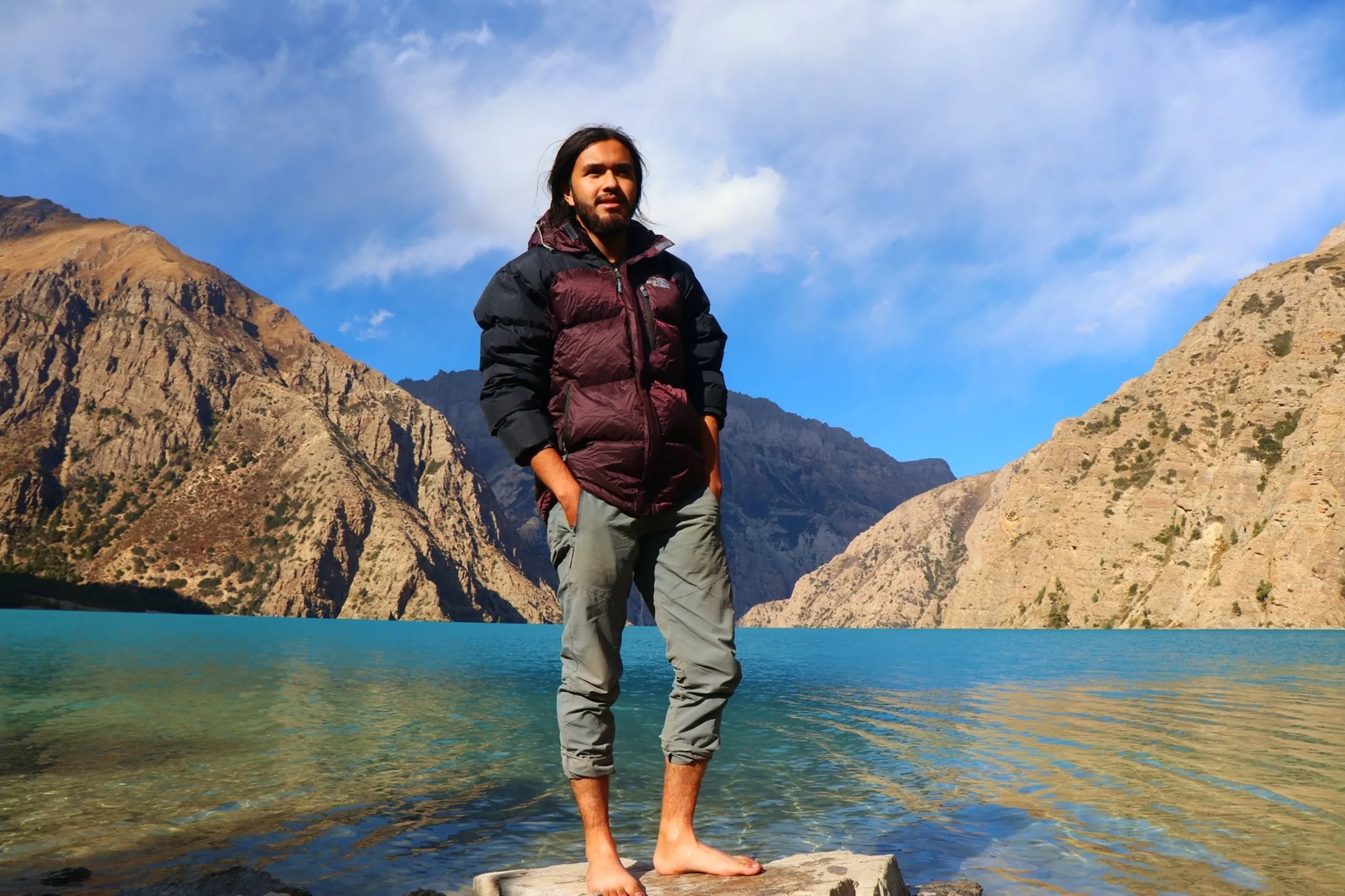 A composed and powerful full-body portrait of a young South Asian male trekker with long dark hair and a beard standing barefoot on a flat boulder at the water's edge of the pristine turquoise Shey Phoksundo Lake, hands tucked into the pockets of his grey trekking trousers, wearing a dark maroon and black two-tone North Face puffer down jacket, looking slightly off-camera with a calm dignified expression, the iconic Shey Phoksundo Lake spreading wide behind him in vivid electric blue-green with dramatic bare rocky grey Himalayan peaks rising symmetrically on both sides under a partly cloudy sky with white clouds drifting overhead, Shey Phoksundo Lake portrait Nepal, male trekker barefoot lake Nepal, North Face Shey Phoksundo trek, Dolpa Nepal lake photography, batch mate lake destination Nepal trek, Himalayan alpine lake Nepal