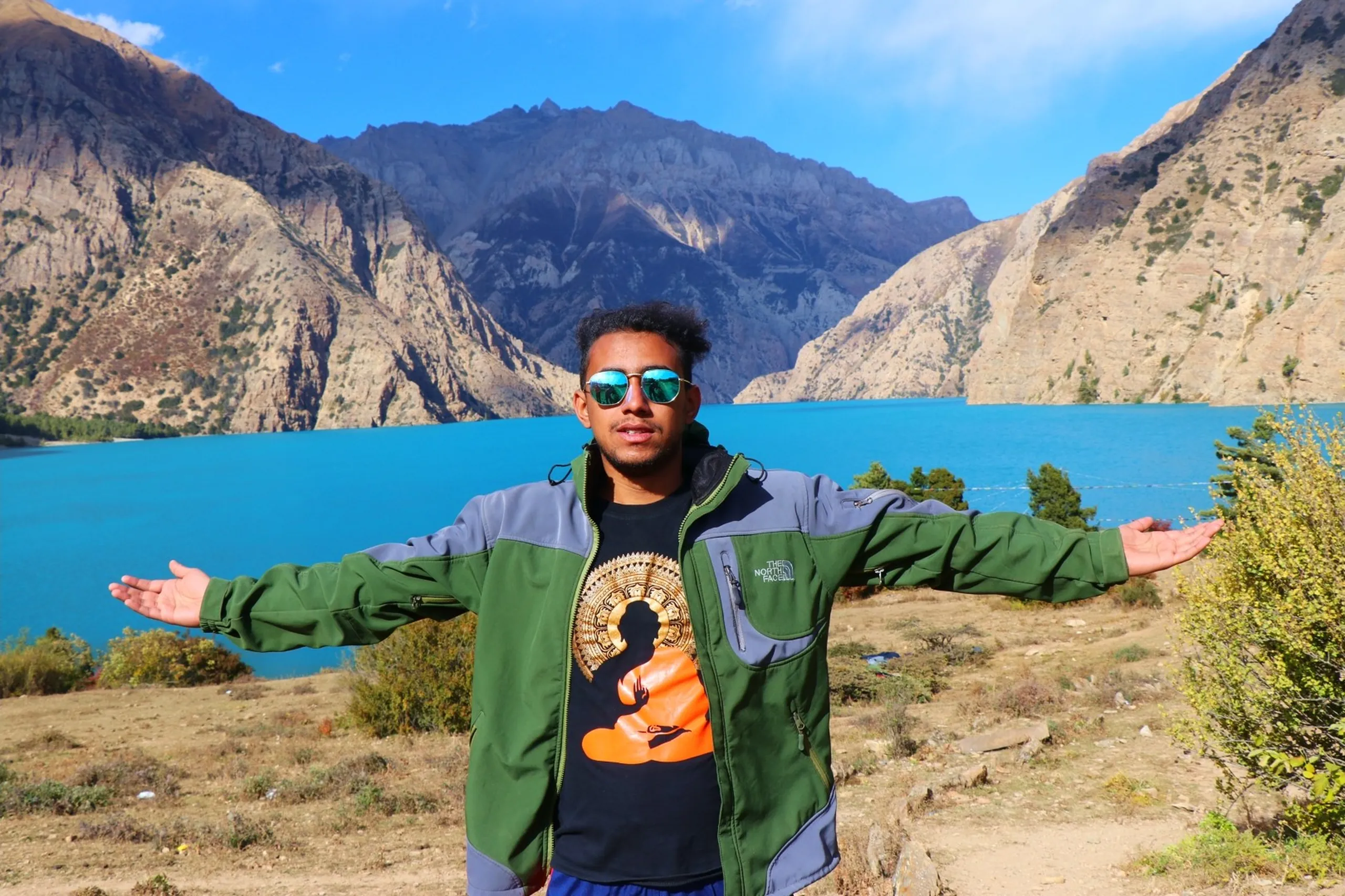 A jubilant and expressive young male trekker with natural curly dark hair and a light beard standing with both arms stretched wide open in a triumphant embrace-the-world pose before the breathtaking Shey Phoksundo Lake, wearing teal-green mirrored aviator sunglasses, an olive green and grey two-tone North Face softshell jacket over a black t-shirt printed with a meditating Buddha illustration and an Aztec/mandala sun design in orange and gold, the iconic vivid turquoise lake filling the entire background between massive barren brown Himalayan canyon walls under a clear blue sky, green shrubs scattered on the rocky slopes nearby, Shey Phoksundo Lake Nepal celebration, arms open Himalayan lake Nepal, North Face trekker Shey Phoksundo, Dolpa Nepal trek milestone celebration, batch mate different college trek Nepal, Shey Phoksundo National Park achievement