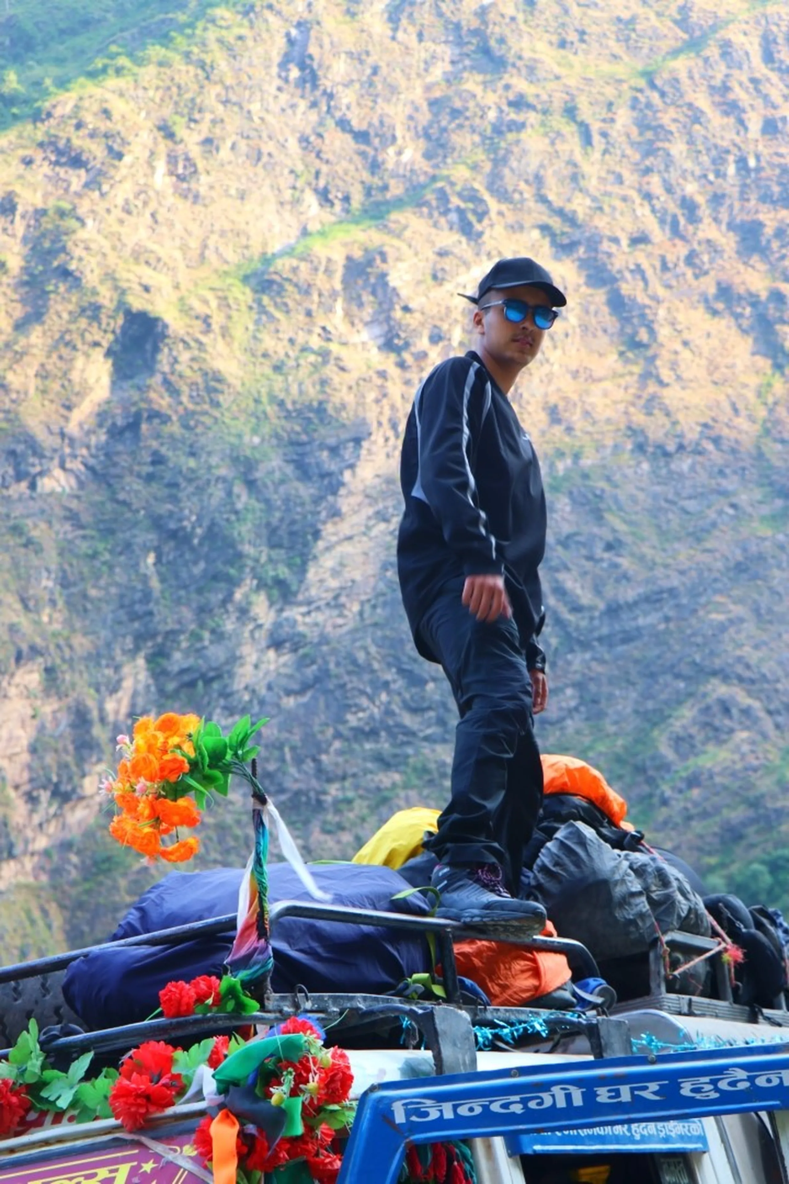 A young South Asian male trekker standing boldly atop the packed luggage roof of a brightly flower-decorated Nepali Bolero jeep, dressed entirely in black tracksuit jacket and pants, wearing a black cap and blue mirrored round sunglasses, striking a confident pose with one foot raised, surrounded by a chaotic array of backpacks, sleeping bags, and colorful artificial flowers in orange and red against a dramatic steep rocky Himalayan cliff face backdrop, Nepali text on jeep sign reads "??????? ?? ?????", rooftop jeep adventure Nepal, extreme travel Himalaya, Nepal trekking youth, Shey Phoksundo National Park journey, daring travel photo Nepal