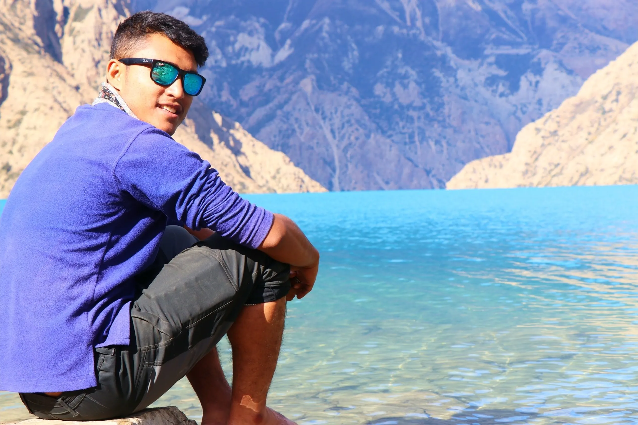 A relaxed and photogenic portrait of a young South Asian male trekker sitting sideways on a low flat stone at the edge of the crystalline Shey Phoksundo Lake, looking back over his shoulder toward the camera with a quiet confident half-smile, wearing a bright cobalt blue long-sleeve fleece top, dark grey cargo shorts and a printed scarf around his neck, with large teal mirrored Ray-Ban wayfarer sunglasses, the impossibly clear shallow turquoise lake water shimmering behind him with perfect reflections, massive barren grey rocky Himalayan canyon walls rising steeply and dramatically on both sides of the lake under a soft blue sky, Shey Phoksundo Lake Nepal sitting portrait, class topper Nepal trek, relaxed lake side trekker Nepal, Dolpa Nepal alpine lake photography, Shey Phoksundo Lake crystal water Nepal, batch mate same college trek achievement Nepal