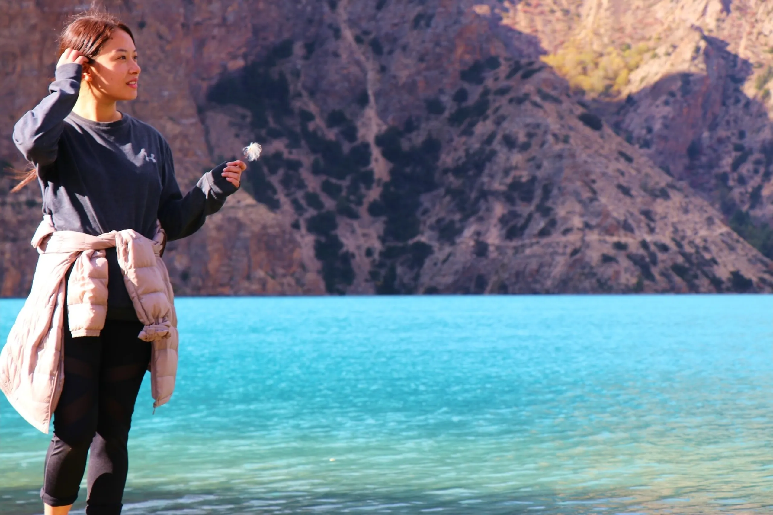 A graceful and serene portrait of a young East Asian-featured South Asian woman standing sideways at the glassy edge of Shey Phoksundo Lake, one hand gently raised to brush wind-blown hair away from her face while the other delicately holds a dried dandelion seedhead, wearing a dark charcoal Under Armour long-sleeve sweatshirt with a beige puffer jacket tied loosely around her waist and fitted black athletic leggings, gazing softly to one side with a quiet smile, the vivid electric turquoise lake water shimmering behind her and a massive barren dark rocky Himalayan cliff face rising steeply in the background, female trekker Shey Phoksundo Lake Nepal, batch junior college friend Nepal trek, dandelion Himalayan lake photography, serene woman Shey Phoksundo, Nepal high altitude lake portrait female, Dolpa Nepal women trek
