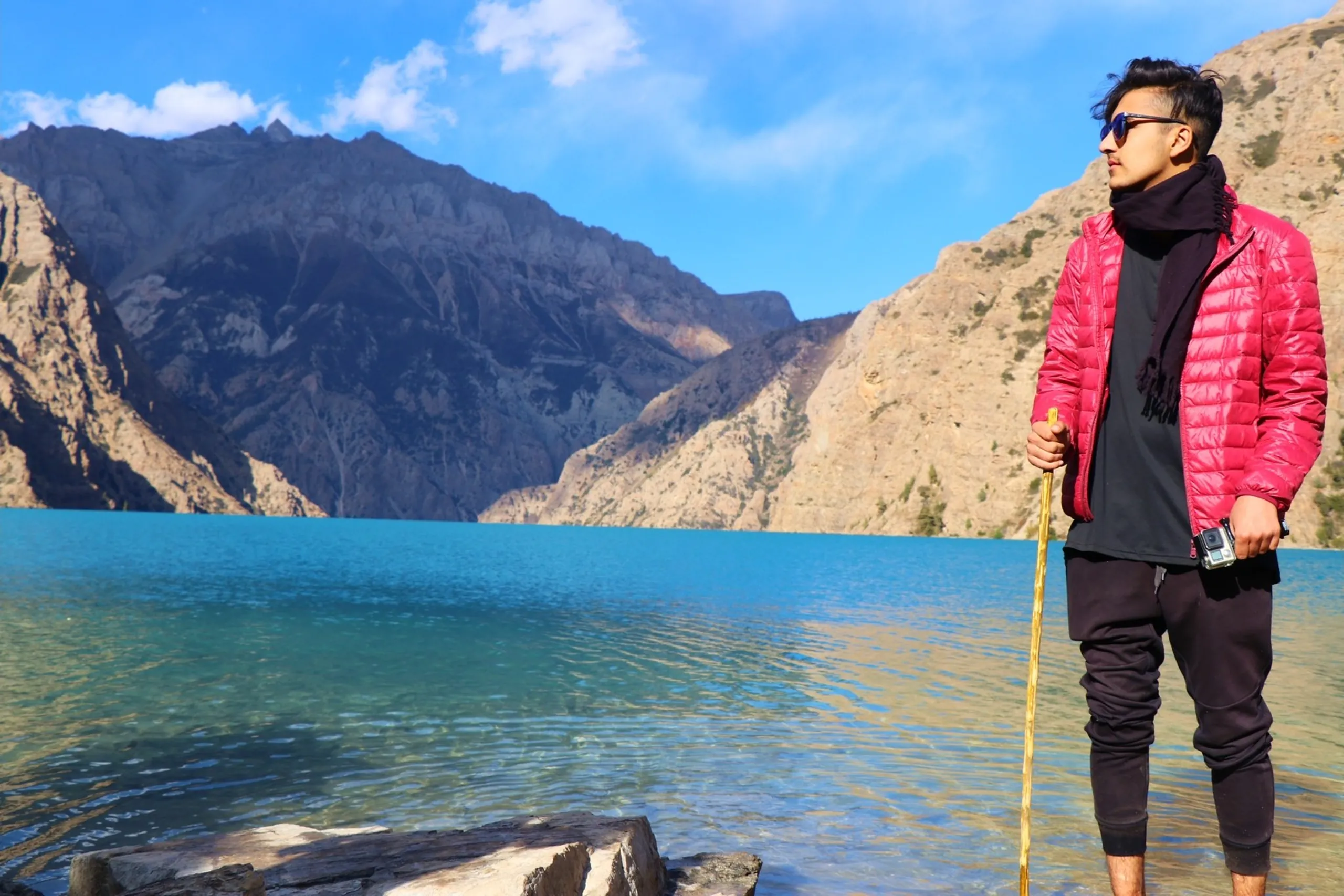 A dramatically composed portrait of a young South Asian male trekker standing at the water's edge of the turquoise Shey Phoksundo Lake, holding a golden bamboo walking stick in one hand and a compact GoPro action camera in the other, wearing a vivid cherry red puffer down jacket over a black hoodie with a black scarf wrapped around his neck and black jogger trousers, blue mirrored sunglasses reflecting the surroundings, gazing confidently to the right across the lake, the iconic electric blue-green lake spreading wide between towering barren grey Himalayan peaks and a few sparse green pine trees under a partly cloudy blue sky, driftwood logs at his feet, Shey Phoksundo Lake Nepal GoPro trekking, red jacket Himalayan alpine lake Nepal, Dolpa trek destination reached, batch junior college trek Nepal, adventure photography Nepal Shey Phoksundo, most beautiful lake Nepal