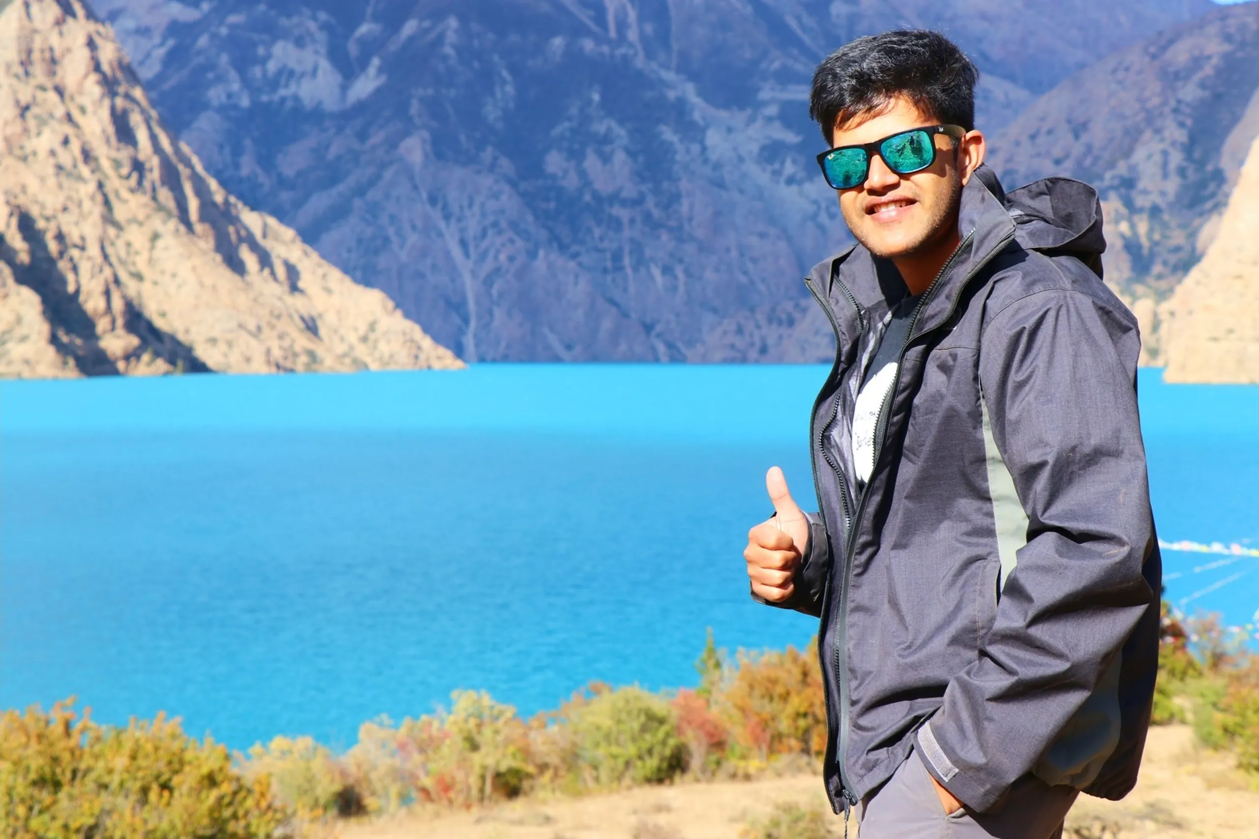 A proud and cheerful young South Asian male trekker giving a thumbs-up at the camera with a bright smile, wearing large dark-tinted teal mirrored rectangular sunglasses and a grey two-tone hooded softshell trekking jacket with a circular badge patch on the chest, standing before the most vivid view of Shey Phoksundo Lake - an impossibly bright electric blue alpine lake spanning the width of the frame between dramatic barren Himalayan rocky ridgelines dotted with yellow-orange autumn shrubs under a brilliant clear blue sky, thumbs up Nepal trek achievement, Shey Phoksundo Lake class topper trekker Nepal, same batch college trek milestone, Dolpa Nepal trek success, Nepal alpine lake thumbs up, Shey Phoksundo National Park achievement trek