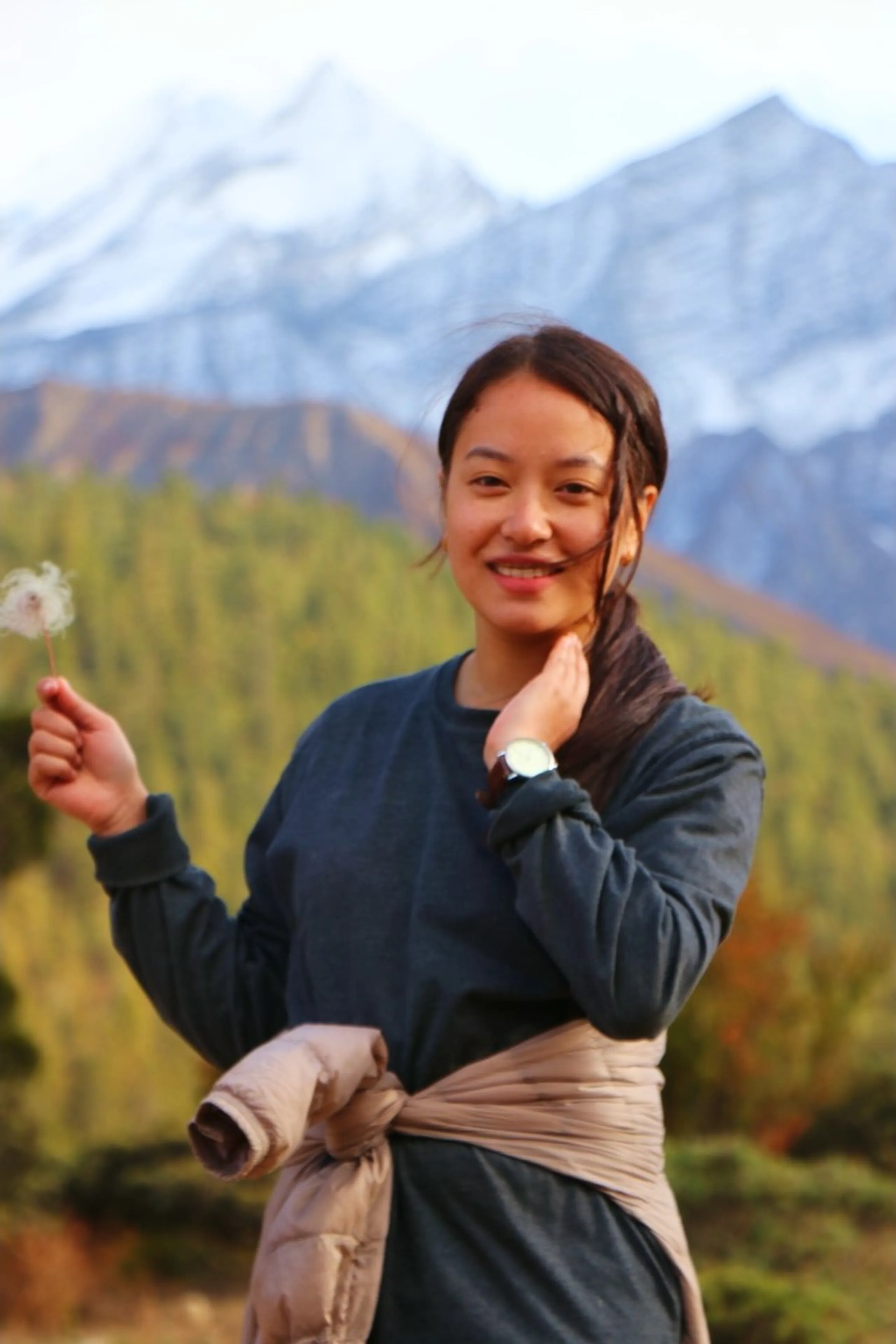 A beautiful close-up portrait of a young East Asian-featured South Asian woman holding a dried dandelion seedhead in her left hand while the other hand touches her neck gently, smiling warmly and directly at the camera with a naturally radiant expression, wearing a dark forest green long-sleeve sweatshirt with a beige puffer jacket tied around her waist and a silver wristwatch, her dark hair pulled back loosely with a few wispy strands framing her face, a stunning soft-focus background of snow-dusted Himalayan peaks, golden-orange autumn forest and green pine trees under an overcast white sky, Nepal female trekker portrait, natural Himalayan beauty Nepal, woman with dandelion Nepal mountains, college batch junior trek Nepal, Shey Phoksundo trek return journey, autumn mountains Nepal Dolpa
