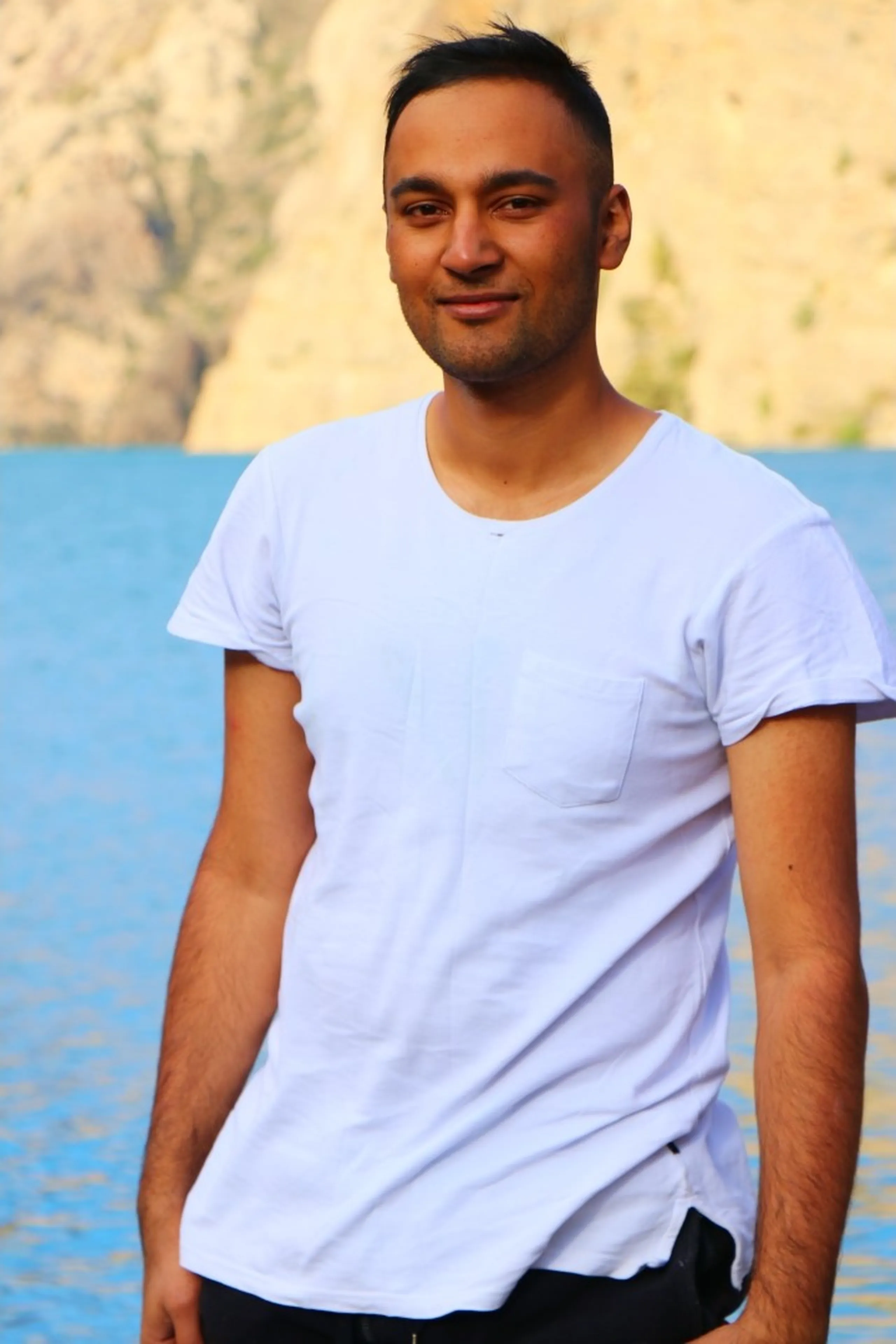 A warm close-up portrait of a young South Asian male with a buzz cut and a gentle genuine smile, wearing a plain clean white round-neck pocket t-shirt, photographed against the blurred vivid turquoise waters of Shey Phoksundo Lake with a barren rocky Himalayan cliff face behind, the blue lake creating a dreamy color-washed bokeh background, casual trekker portrait Nepal Shey Phoksundo, Dolpa trek friend group Nepal, outsider joined trek Nepal, Nepal mountain portrait photography, Shey Phoksundo Lake background portrait, Himalayan trek friendship Nepal