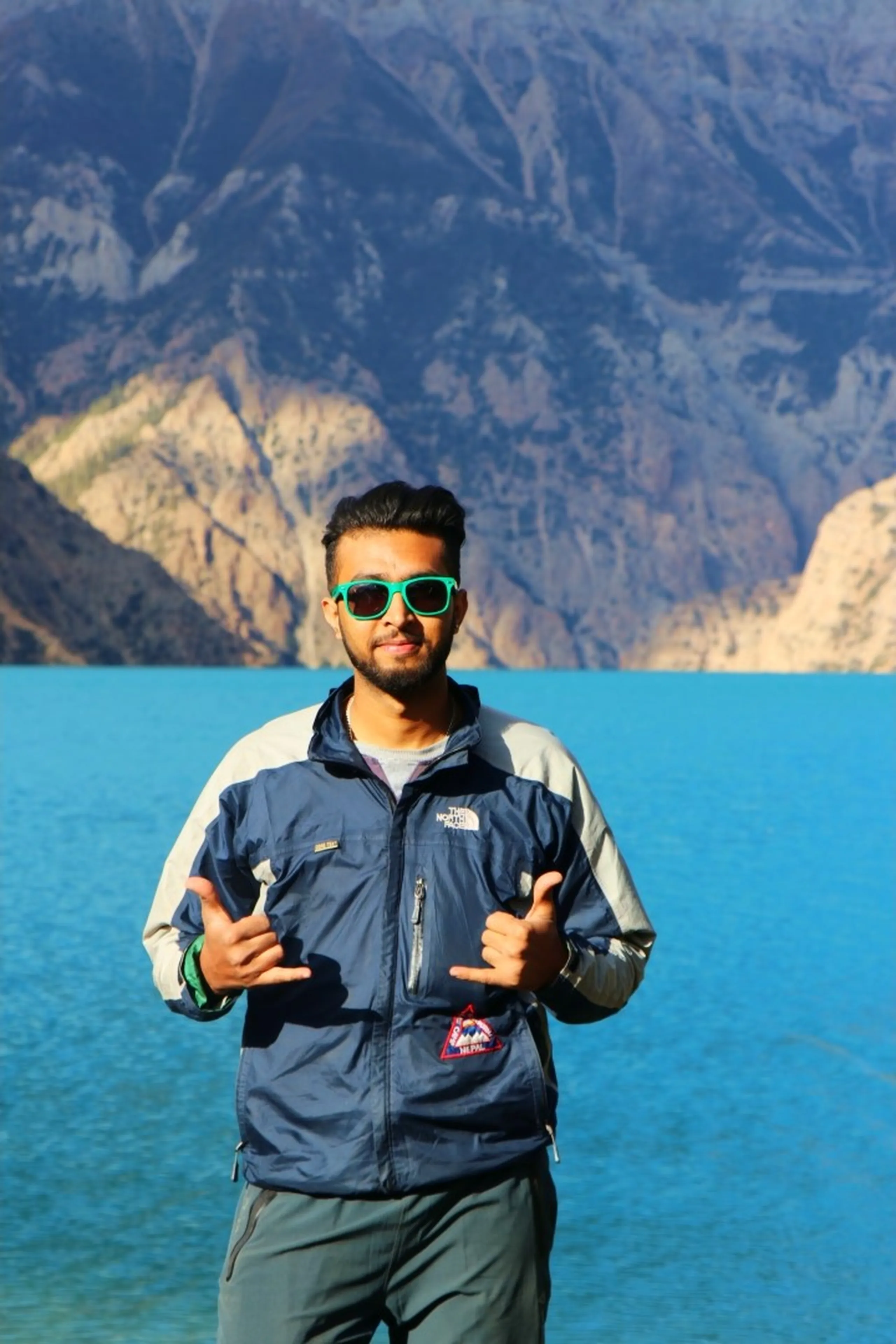 A confident and stylish young South Asian male trekker posing with both hands making a casual double shaka or gun-finger gesture directly at the camera with an assured expression, wearing bold lime green-framed wayfarer sunglasses, a two-tone navy and silver-grey North Face windbreaker jacket with a circular trek badge patch on the chest, and grey trekking trousers, standing directly before the vivid turquoise blue expanse of Shey Phoksundo Lake with massive arid Himalayan rocky peaks towering on both sides in the background, Nepal trek photographer Shey Phoksundo, college same batch photographer trekker Nepal, North Face gear Dolpa Nepal trek, trek photographer Shey Phoksundo Lake Nepal, adventure photographer trekking Nepal Himalaya, turquoise lake Nepal photographer