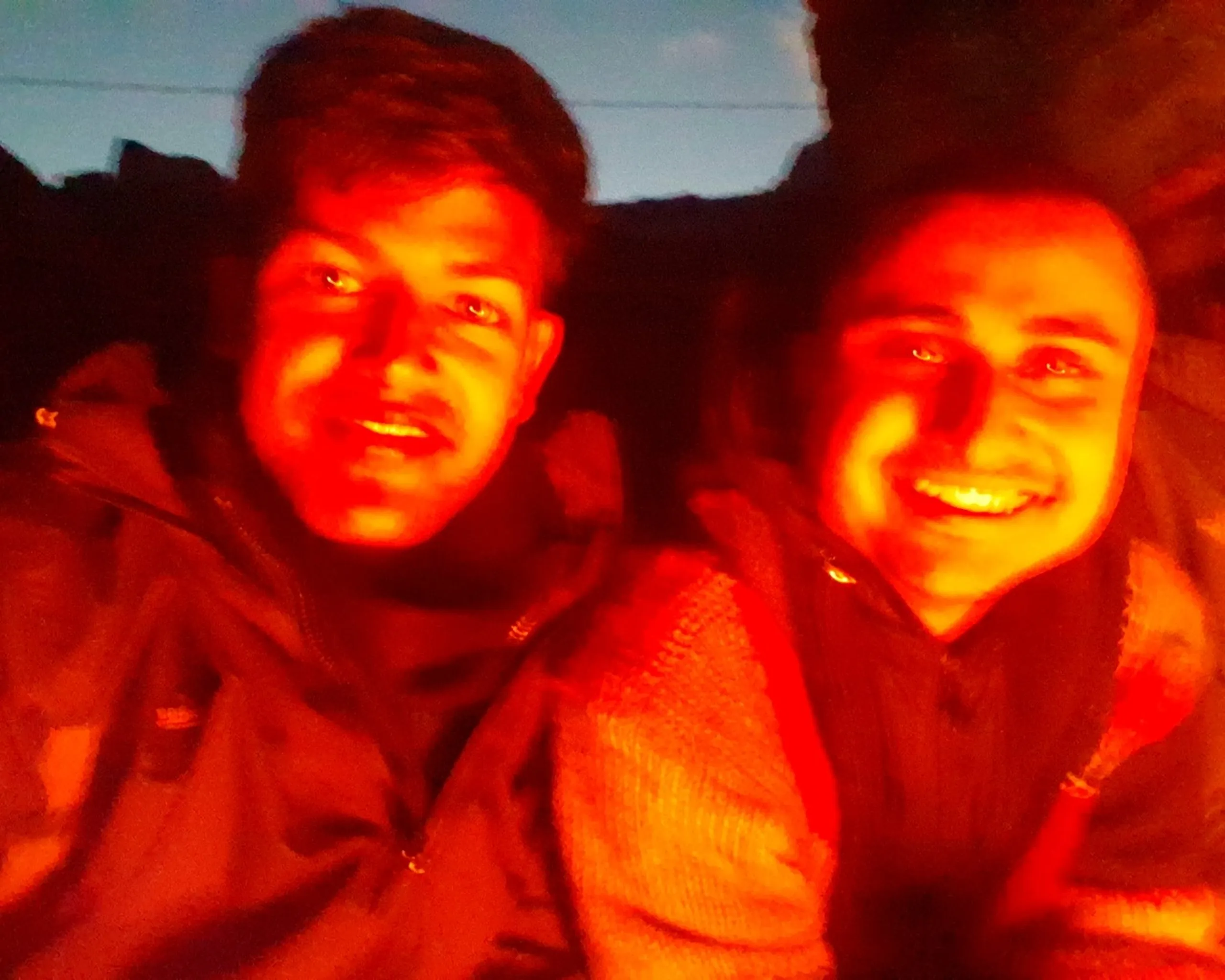 A warmly lit low-light nighttime campfire selfie of two young South Asian male trekkers illuminated entirely by the orange glow of a wood fire or bonfire, both grinning broadly with shining eyes - one on the left in a dark jacket looking directly into the lens, the other on the right in a multicolored knitted sweater with equally bright warm smile, the dark rocky Himalayan silhouettes barely visible against a deep blue twilight sky behind them, an intimate and joyful moment captured during the overnight stay at Sherpa Hotel near Shey Phoksundo in freezing night temperatures, campfire night Nepal Himalaya trek, cold night campfire Dolpa Nepal, batch mates bonding Nepal trek, trekking friends campfire Shey Phoksundo, Sherpa hotel Nepal wood fire night, winter camping Nepal mountain