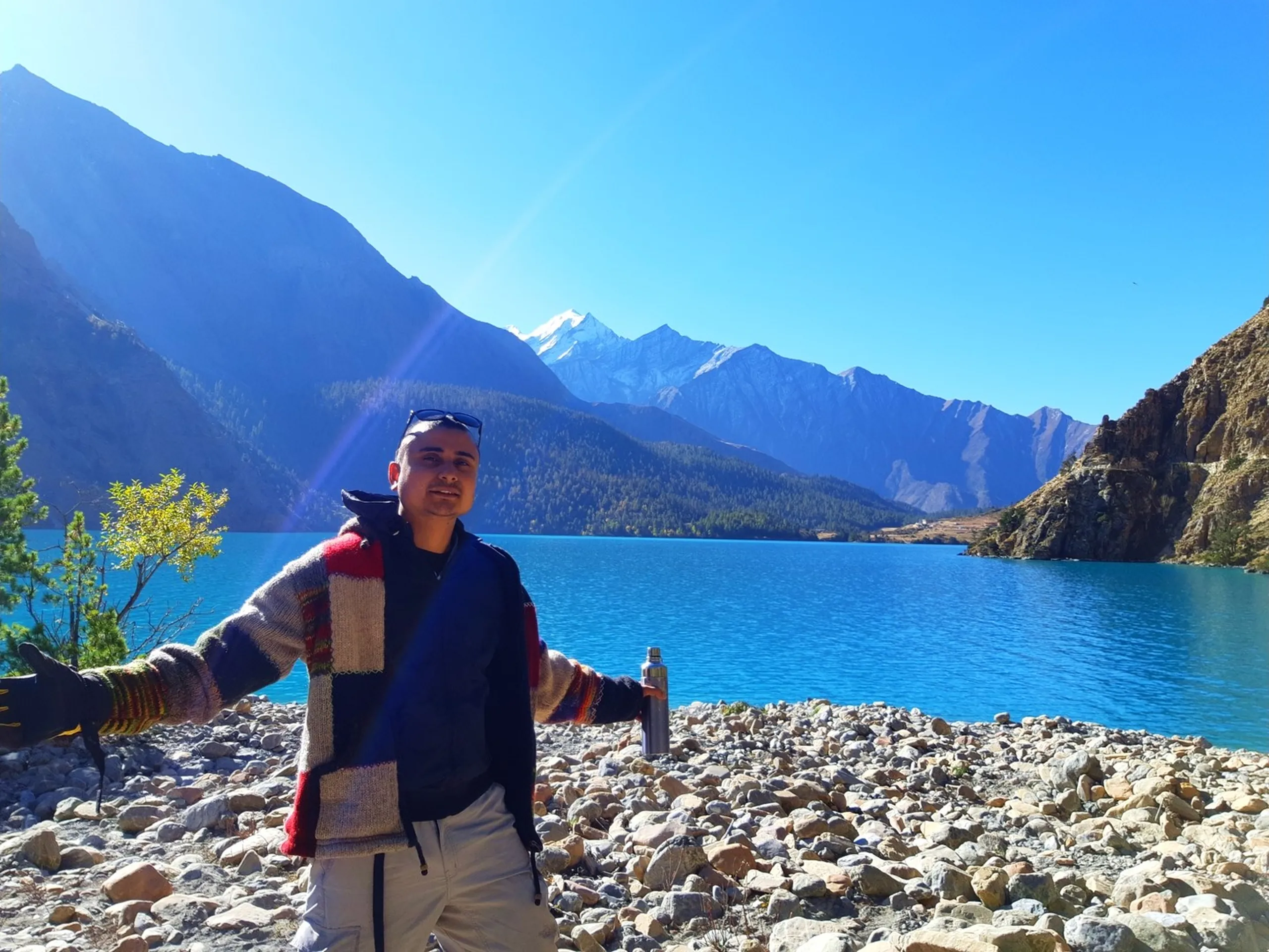 A wide-angle landscape-format portrait of a young South Asian male trekker standing with arms spread open in a relaxed welcoming pose on a rocky pebbly shoreline of the magnificent Shey Phoksundo Lake, wearing a colorful traditional Nepali handwoven striped wool sweater in bold red, navy, beige and multicolor horizontal stripes layered over a dark blue jacket, light grey trekking trousers and dark sunglasses pushed up on his forehead, holding a stainless steel thermos flask in one hand, a small green tree to the left, the vivid electric blue lake stretching behind him with dense green pine forests on the far shore and a dramatic snow-capped Himalayan peak rising majestically in the distance under a bright sunny blue sky with a visible sun flare, Shey Phoksundo Lake Nepal arms open trekker, traditional Nepali wool sweater hiking, Dolpa trek lake shoreline Nepal, batch mate college friend Shey Phoksundo, Nepal thermos flask mountain lake, most scenic lake Nepal trekking