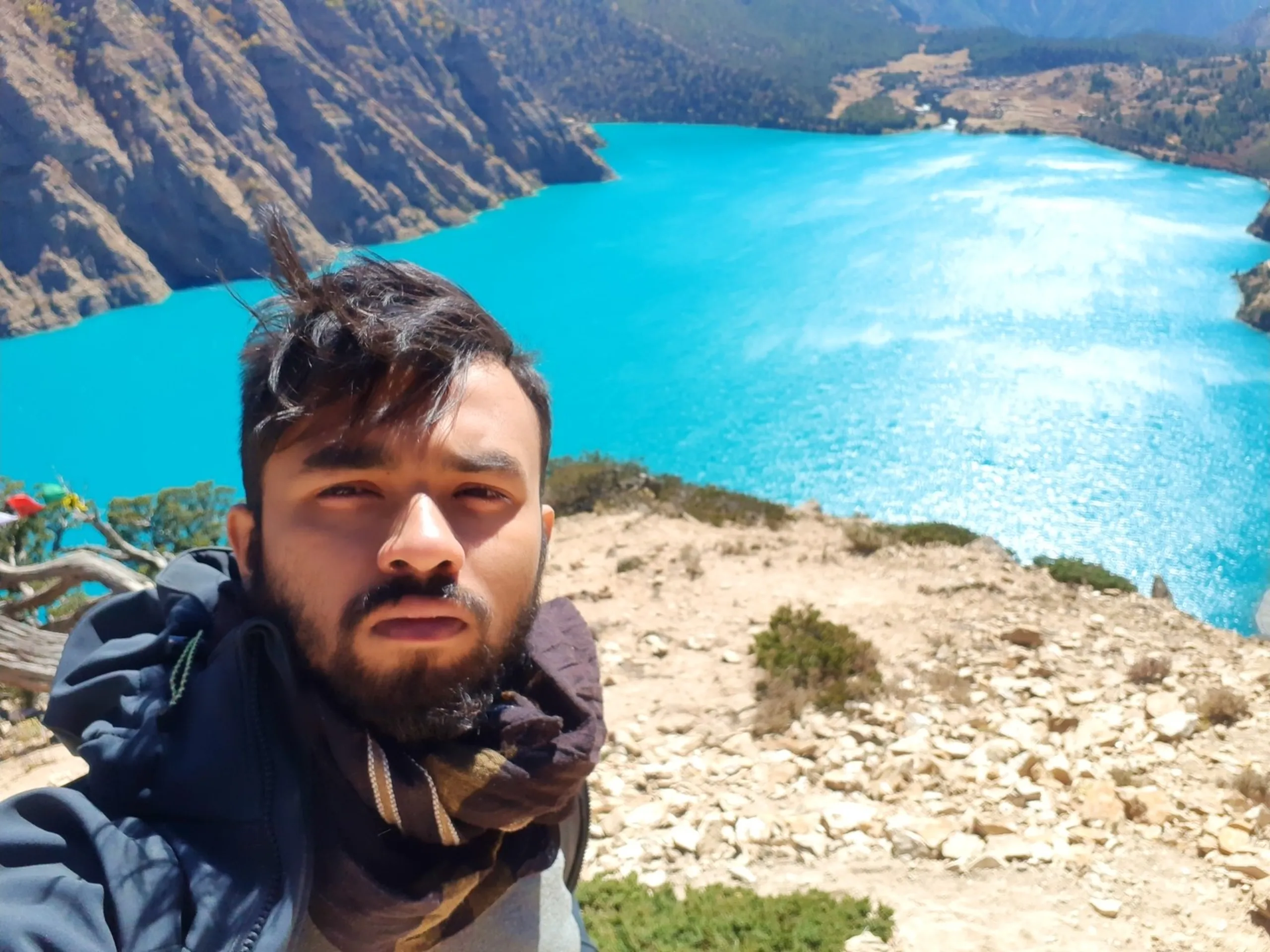 A daring high-altitude selfie taken from a clifftop viewpoint directly above Shey Phoksundo Lake, the trekker's bearded face filling the left foreground of the frame - hair dramatically blown sideways by strong mountain winds, wearing a dark jacket with a scarf wrapped around his neck - while far below and behind him the entire spectacular Shey Phoksundo Lake is visible as a sweeping vivid turquoise oval nestled between barren grey rocky Himalayan canyon walls, a twisted gnarled ancient tree with red prayer flags barely visible at the cliff edge, the scale of the lake made tiny by the dramatic height of the viewpoint, Shey Phoksundo Lake bird's eye view Nepal, clifftop selfie Nepal Himalaya, Dolpa lake aerial view selfie, trekking viewpoint Nepal Shey Phoksundo, highest viewpoint Shey Phoksundo National Park, Nepal adventure photography wind