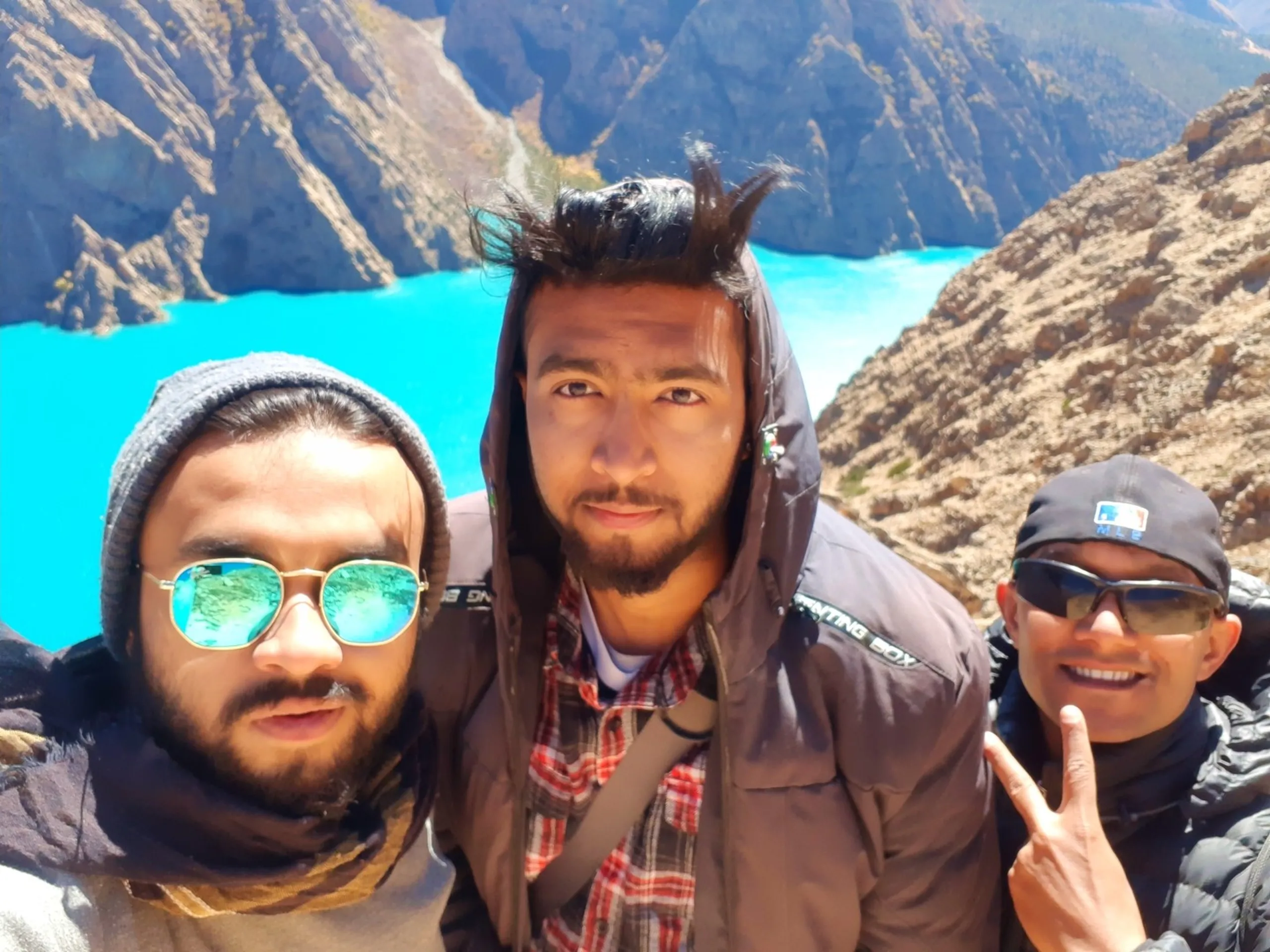 A fun windy three-person selfie taken from a dramatic clifftop viewpoint high above Shey Phoksundo Lake - the person taking the selfie on the left wearing a grey wool beanie and round gold-framed teal mirrored sunglasses with a scarf, the middle trekker with wild windswept hair sticking straight up dramatically from his head wearing a brown "Hunting Box" hooded jacket and red plaid scarf, and a third trekker on the right in a black puffer jacket and MLB-logo cap with dark sunglasses flashing a peace sign - the dazzling turquoise Shey Phoksundo Lake glowing far below between jagged Himalayan rocky ridgelines, Shey Phoksundo viewpoint group selfie Nepal, windy clifftop selfie Dolpa Nepal, Dhiraj Kusal Ramesh Shey Phoksundo Nepal, best viewpoint Nepal trek, trekking trio Shey Phoksundo Lake above, Nepal mountain winds funny selfie