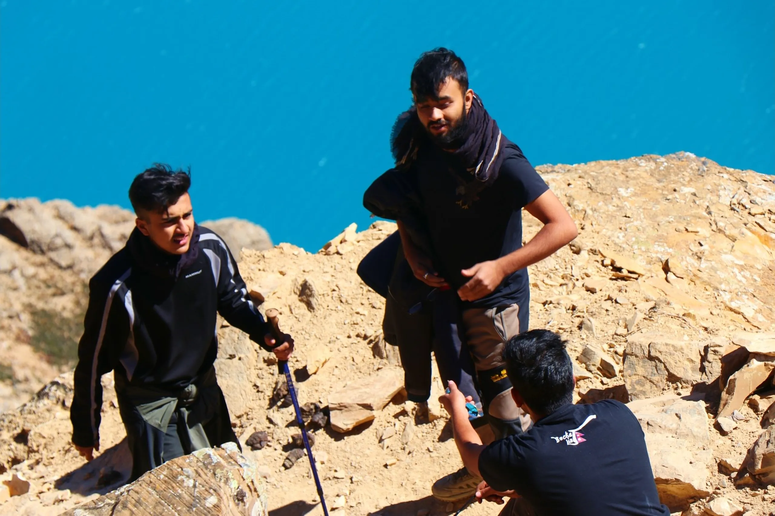 A candid action photograph of three male trekkers navigating a steep rocky scramble on a loose boulder-strewn hillside above the turquoise Shey Phoksundo Lake - one on the left in a black Mammut brand jacket using a trekking pole for balance and squinting against the sun while ascending, a bearded trekker in the center wearing a black t-shirt and scarf with grey cargo pants helping steady himself on a large rock, and a third crouching below him in a black "Bucket List" branded jacket holding a compact camera or video device to document the moment, the vivid blue lake visible in the background behind the rocks, candid trek scramble Nepal Shey Phoksundo cliff, trekkers rocky scramble Dolpa Nepal, batch mates helping each other Nepal mountains, off trail scramble Shey Phoksundo viewpoint, adventure Nepal Himalaya group trek, Bucket List Nepal trek