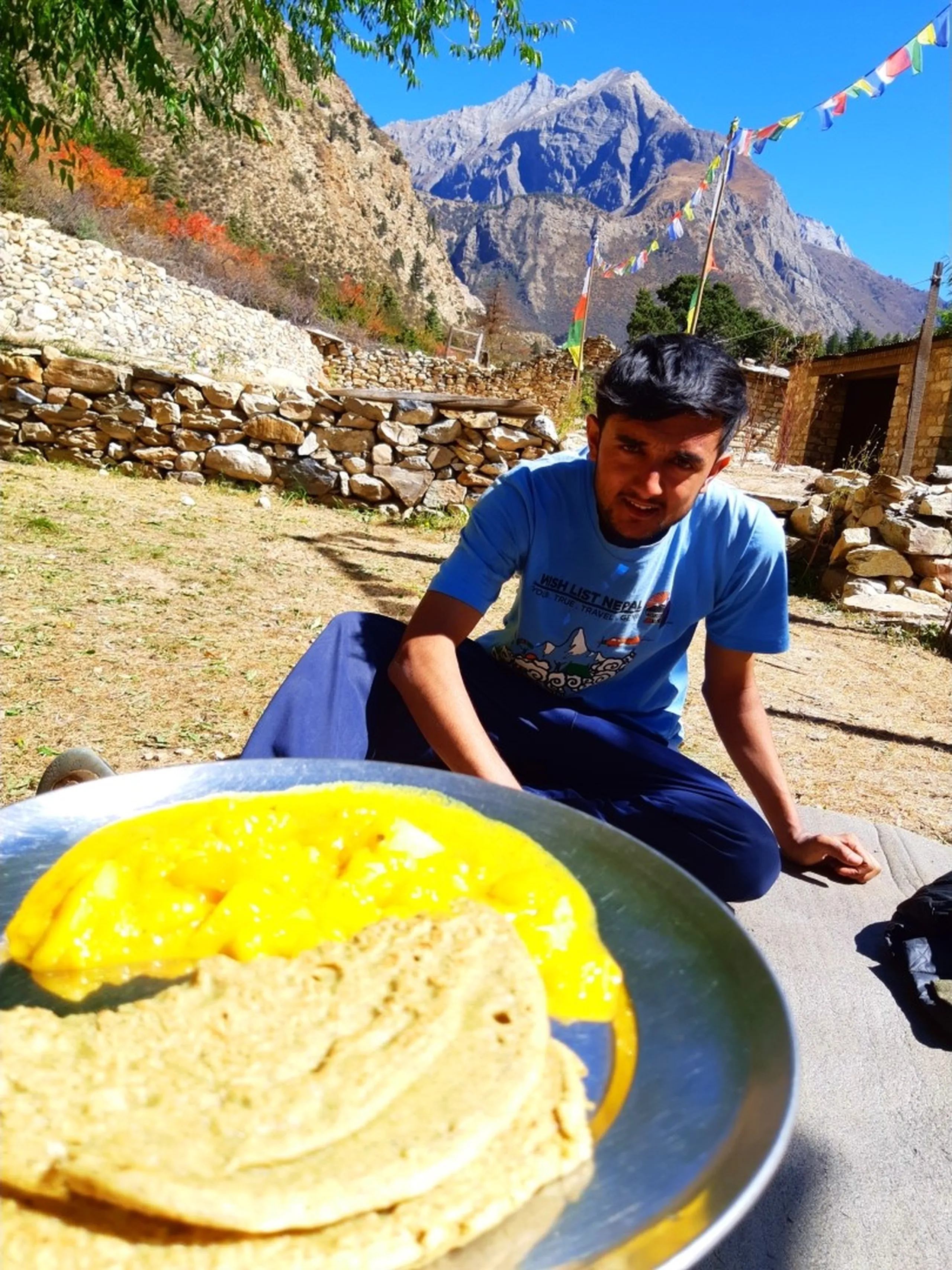 A playful close-up food photography shot showing a round aluminium plate heaped with golden yellow saag aloo (potato curry) and a flat whole wheat roti/chapati served as a typical Nepali trekking lunch at Jharna Hotel, the vibrant yellow curry filling the frame in the foreground while in the background a young South Asian male trekker in a light blue "Wish List Nepal - Your True Travel Genie" branded t-shirt leans over the food with an eager expression, sitting on the ground of a remote high-altitude village courtyard with traditional dry-stone boundary walls, colorful Buddhist prayer flags strung overhead and a dramatic bare Himalayan peak rising under a clear blue sky behind, typical Nepali trek food, dal bhat roti Nepal mountain lunch, Wish List Nepal tour trekking food, Jharna Hotel Dolpa Nepal, authentic Nepal mountain cuisine, trekking meal Nepal Shey Phoksundo