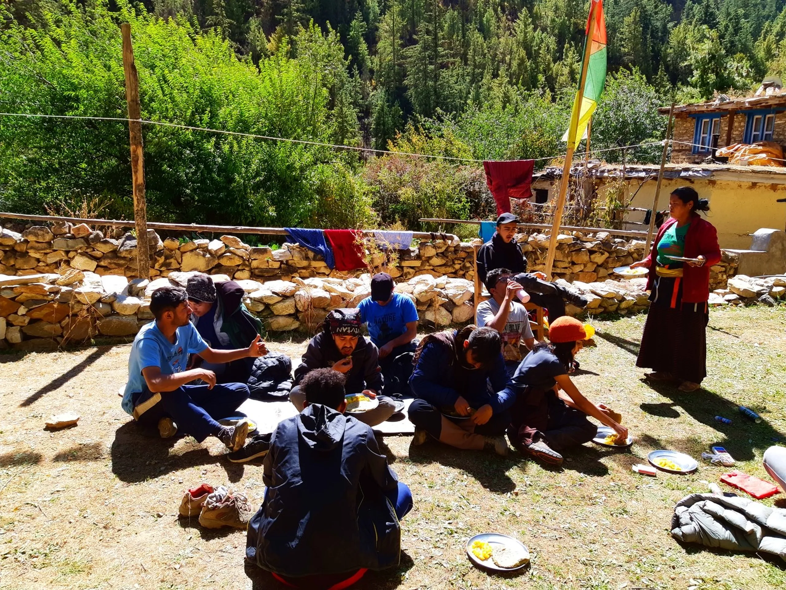 A wide candid group photograph of the entire Shey Phoksundo trekking group seated together in a loose circle on a sunny grass clearing of a remote Nepali mountain village, eating lunch together from round aluminium plates and bowls, wearing a mix of hiking jackets, bandanas, blue t-shirts and dark outerwear - approximately ten people including men and women - surrounded by traditional stone-walled enclosures, a blue-windowed stone house, a tall colorful prayer flag pole (green-orange-red) and green pine forest behind, while a local Nepali woman in traditional red and dark maroon dress and striped apron serves food to the group, the atmosphere radiating genuine warmth and community spirit, Nepal trekking group lunch outdoor, Shey Phoksundo trek team meal, remote Nepal village food together, trekking family Nepal Dolpa, authentic Nepali hospitality mountain trek, group dining Nepal Himalaya