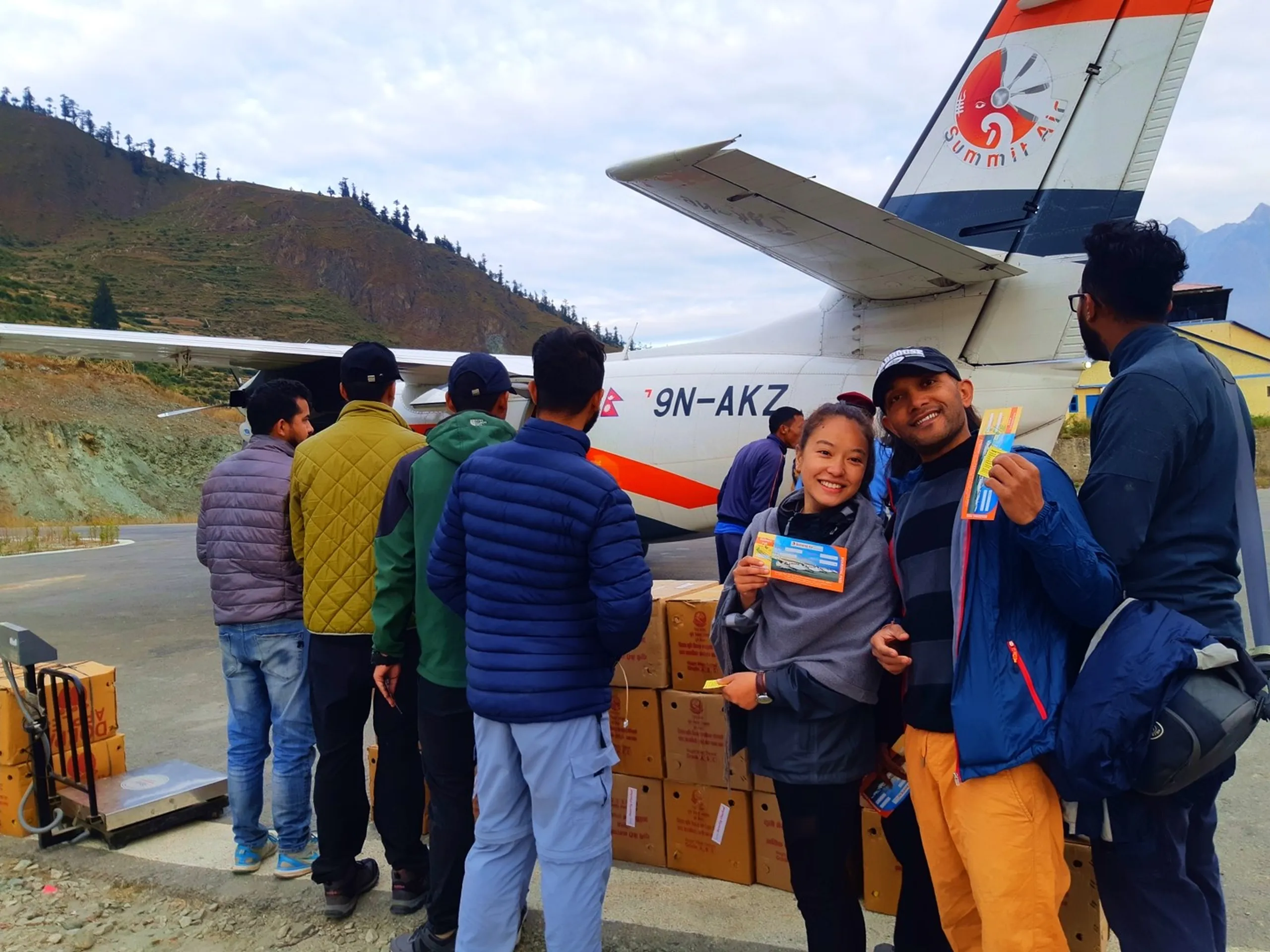 A vibrant airport tarmac scene at a remote Himalayan mountain airstrip in Dolpa, Nepal - a white twin-turboprop Summit Air aircraft with registration 9N-AKZ and the distinctive Summit Air red logo visible on the tail fin is parked while being loaded with cardboard supply boxes, a queue of trekkers and locals in colorful jackets (yellow, navy, olive green) standing in line to board - while in the foreground a young East Asian-featured South Asian woman wearing a dark grey fleece jacket and a black cap holds up an orange-and-blue printed boarding pass ticket and smiles brightly beside a young bearded male trekker in orange trousers also holding his boarding ticket with a broad grin, Summit Air Nepal Dolpa airport flight, mountain airstrip Nepal, Shey Phoksundo trek return flight Nepalgunj, Dolpa Nepal airport boarding, Summit Air Nepal 9N-AKZ, remote Himalayan airstrip Nepal travel