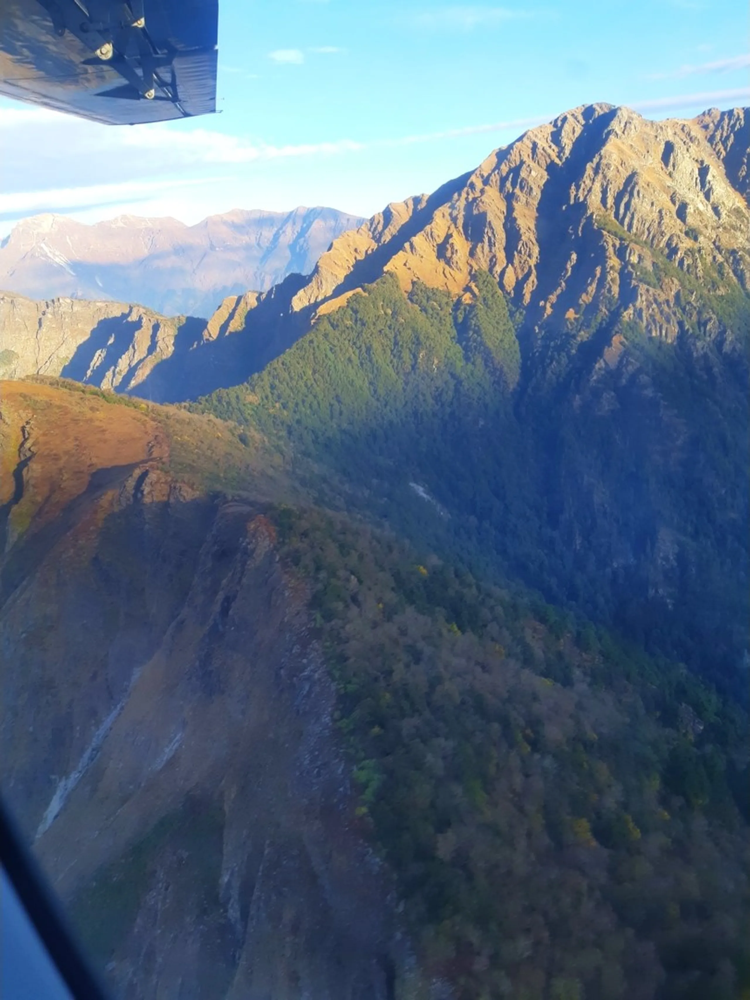 A breathtaking aerial window-seat view photographed from inside a small propeller aircraft flying through the Himalayan mountain ranges of the Dolpa district of Nepal, the aircraft's dark engine and wing tip visible in the upper left corner of the frame, looking out over a dramatic layered landscape of towering ridgelines covered in patches of golden-brown autumn vegetation and dense dark green pine forest dropping steeply into deep shadowed gorges and valleys, snow-capped distant peaks barely visible through atmospheric haze on the horizon under a partly cloudy blue sky, capturing the rugged remote and inaccessible terrain of the Dolpa region that makes this one of Nepal's most remote and challenging treks, aerial view Dolpa Nepal mountains, Himalayan aerial photography Nepal, Summit Air flight Dolpa Nepalgunj Nepal, remote mountain flight Nepal, Shey Phoksundo trek aerial return, flying over Nepal Himalaya