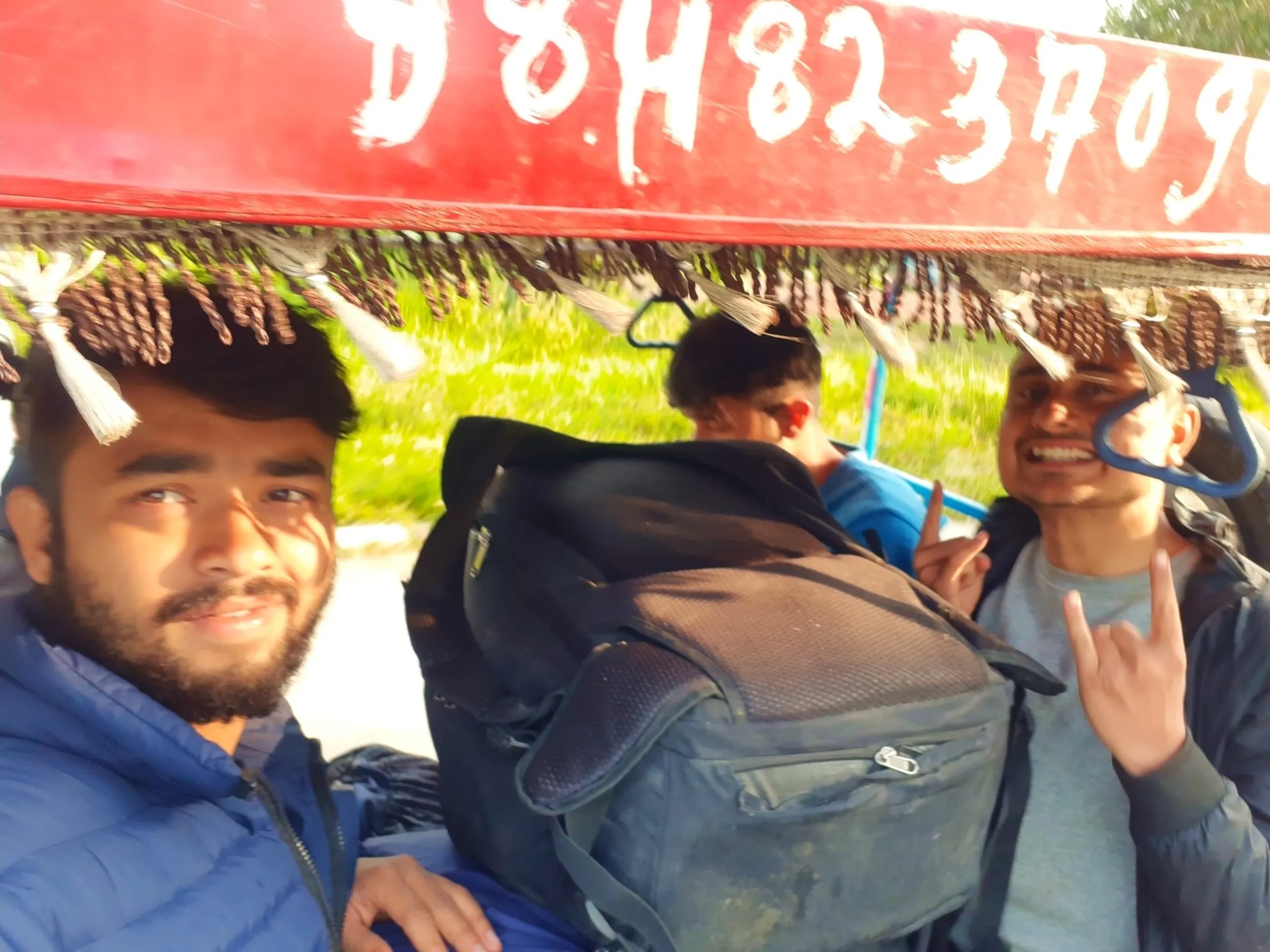 A candid blurry motion selfie of two young South Asian male trekkers sitting inside a moving decorated Nepali rickshaw or tempo vehicle, the red canopy of the vehicle visible above them with tasseled fringe trim and a hand-painted phone number "9848237096" on the red signboard, one on the left in a navy blue puffer jacket with a trimmed beard looking softly at the lens, a backpack between them belonging to a third person visible from behind in the middle, while the trekker on the right in a light grey sweater with short buzz cut beams a wide grin while flashing a peace sign and rock-and-roll hand gesture, green roadside vegetation rushing past outside, Nepal tempo ride Nepalgunj, trekkers celebrating return Nepal, batch mates Nepalgunj Nepal, rickshaw selfie Nepal lowlands, post trek celebration Nepal, after Dolpa trek Nepal journey back