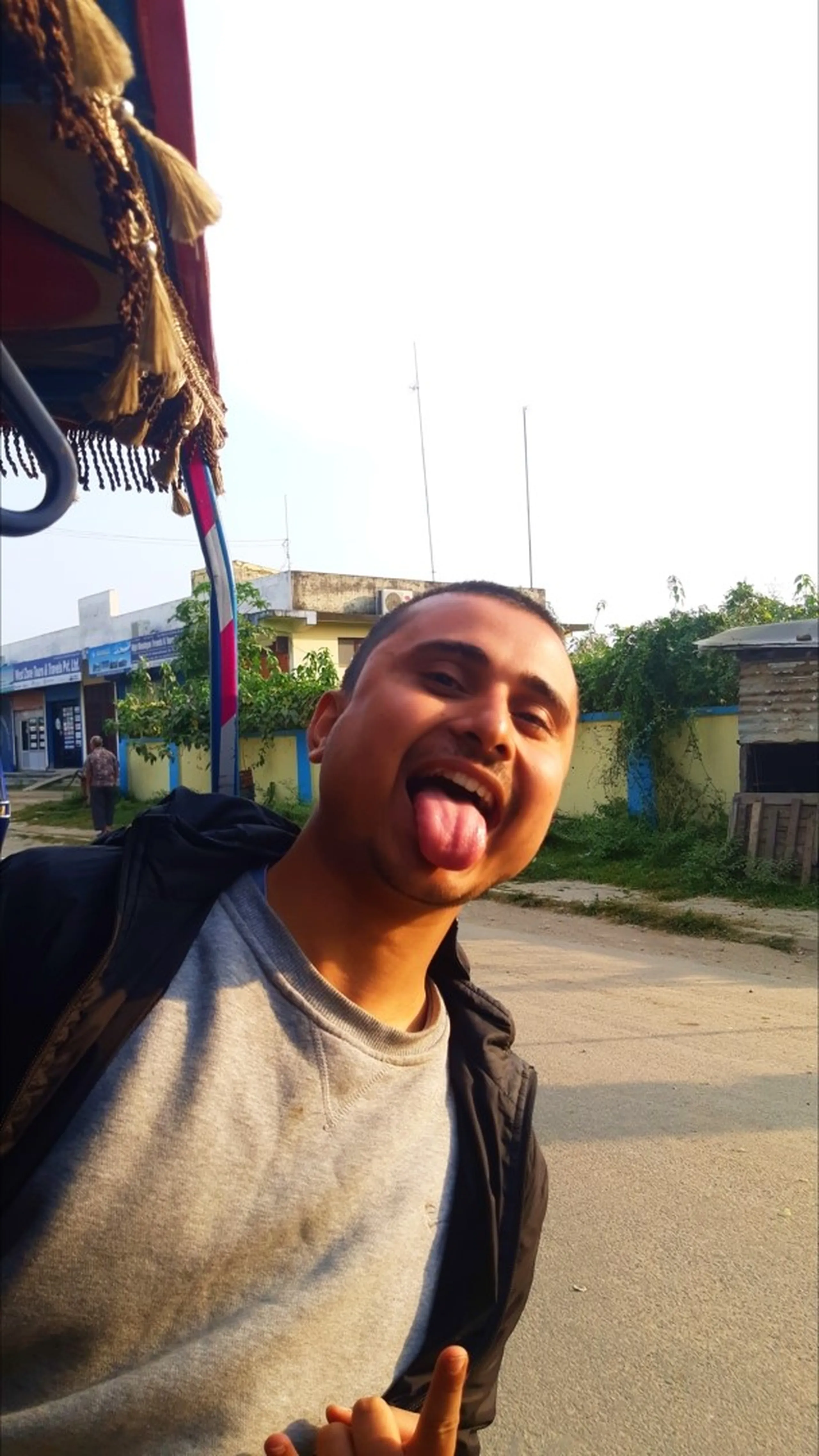 A hilarious and candid close-up selfie of a young South Asian male trekker with a buzz cut sticking his tongue fully out of his mouth in a wild playful expression directly at the camera, wearing a light grey crewneck sweatshirt under a dark outer jacket, photographed under the red tasseled canopy of a Nepali rickshaw or tempo with blurred low-rise Nepalgunj buildings, antenna towers and green trees visible in the out-of-focus background, the raw unfiltered joy of completing the Shey Phoksundo trek captured in one goofy unforgettable expression, funny trekker Nepal post trek, Nepalgunj Nepal celebration selfie, batch mates fun Nepal return journey, goofy trekking celebration Nepal, Dolpa trek completion Nepal humor, lighthearted Nepal travel blog photo