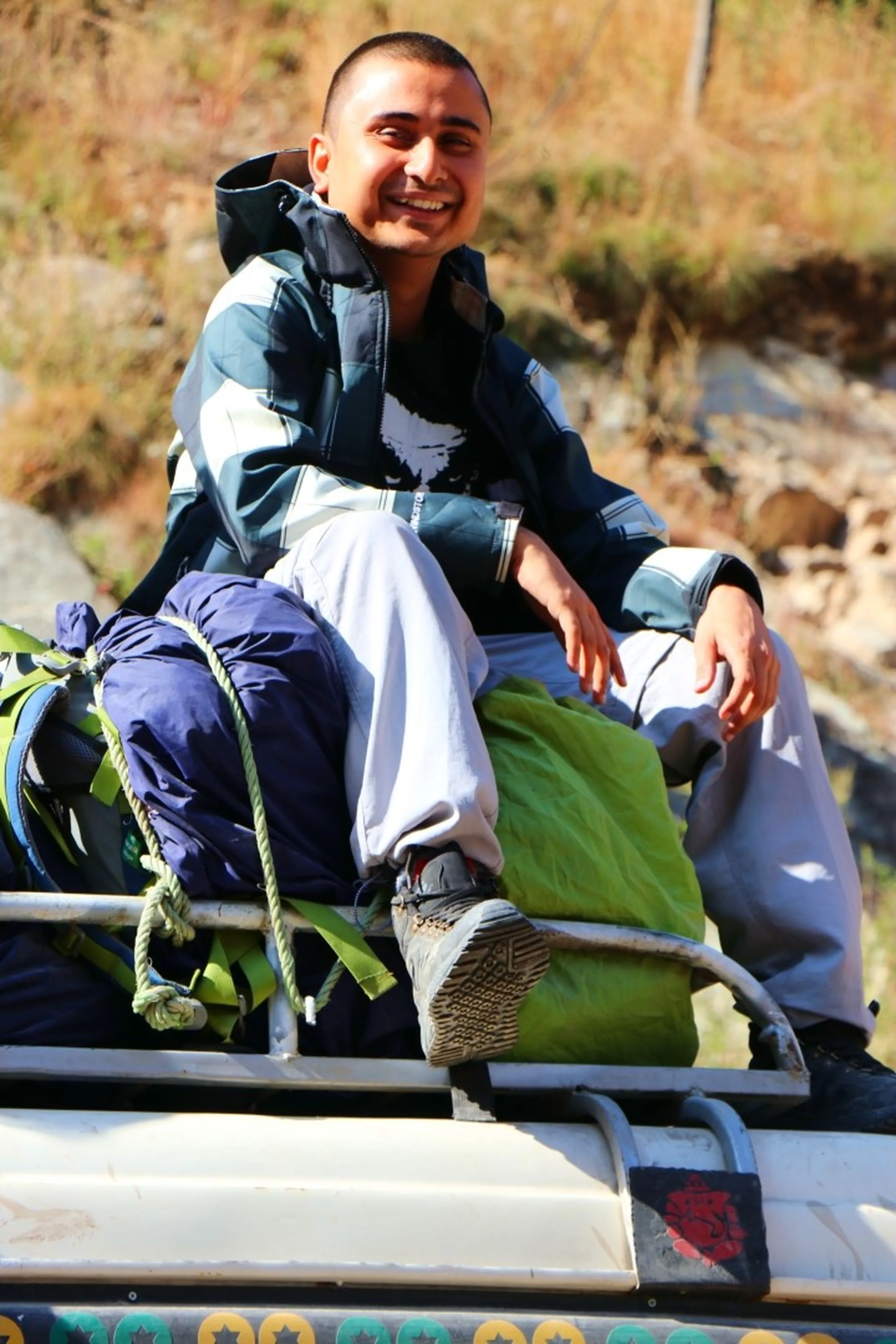 A cheerful young South Asian male trekker with a buzz cut and a wide beaming smile sitting on top of a jeep roof rack loaded with tightly roped blue and green sleeping bags and backpacks, wearing a navy blue and grey North Face-style puffer jacket and light grey trekking trousers with rugged hiking boots, sunny rocky hillside with dry golden grass in the background, rooftop jeep travel Nepal, adventure trekking Himalaya, happy trekker Nepal mountains, Shey Phoksundo trek preparation, budget travel Nepal backpacker