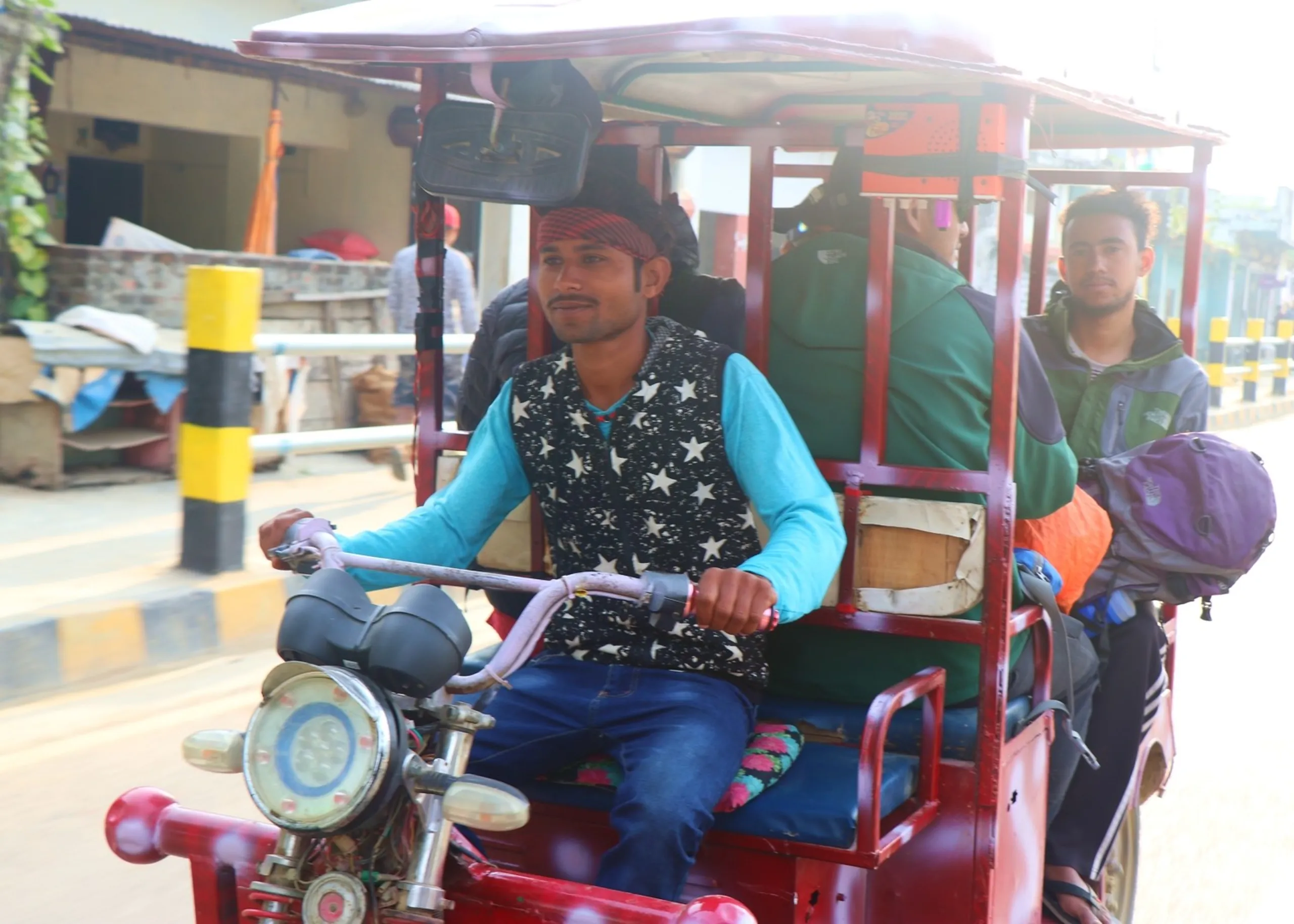 A candid street-level action photograph taken from inside a vehicle showing a young Nepali man in a teal long-sleeve top, black star-printed vest and jeans wearing a red bandana headband confidently driving a red electric rickshaw or e-rickshaw through a busy Nepalgunj street, gripping the handlebars with a relaxed half-smile, while in the back seat a trekker in a green North Face vest carries a large purple and orange backpack, other trekkers partially visible in the back of the vehicle, bright overexposed morning sunlight flooding the scene, yellow and black traffic bollards, local buildings and shops visible along the Nepalgunj street, Nepal e-rickshaw local transport, Nepalgunj street travel Nepal, trekkers local transport Nepal lowlands, Nepal electric rickshaw adventure, post-Dolpa trek Nepalgunj return, real Nepal street life travel photography