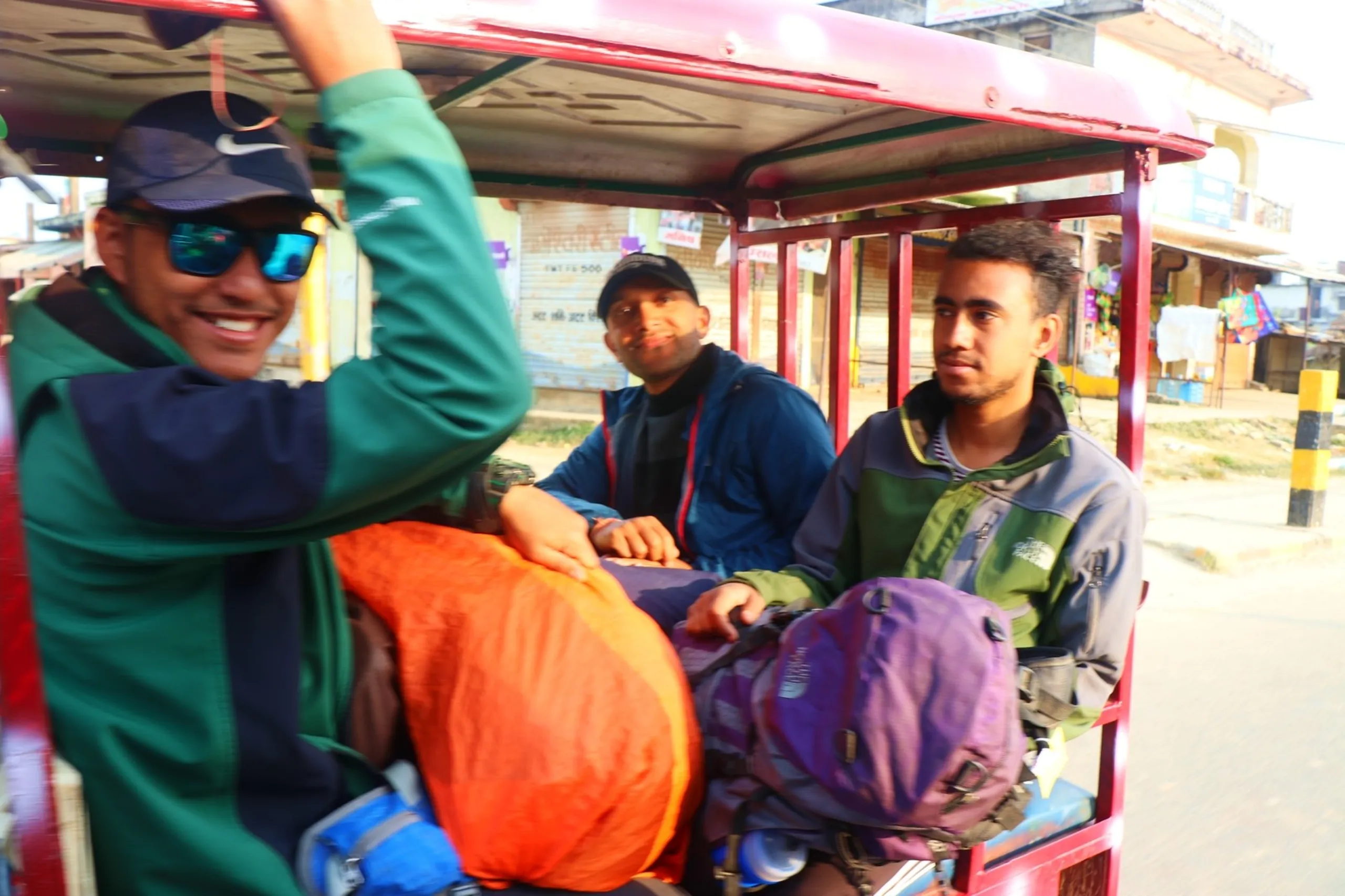 A candid action shot inside a moving red Nepali electric rickshaw or tempo crammed with trekking gear, capturing three young South Asian male trekkers in high spirits on the streets of Nepalgunj - one on the left in a green and navy jacket wearing teal mirrored sunglasses and a black Nike cap grins broadly while holding the overhead bar with one arm raised, a batch mate in a black jacket and black cap smiles warmly in the middle, and a third in an olive green North Face vest jacket sits quietly on the right with a large purple backpack between them, an orange sleeping bag roll and blue bag visible on laps, blurred Nepalgunj storefronts and shuttered shops rushing past outside, Nepalgunj Nepal e-rickshaw post trek, trekkers return journey Nepal lowlands, batch mates Nepalgunj celebration ride, Shey Phoksundo trek return Nepalgunj, Nepal local transport trekkers, outsider joined group trek Nepal