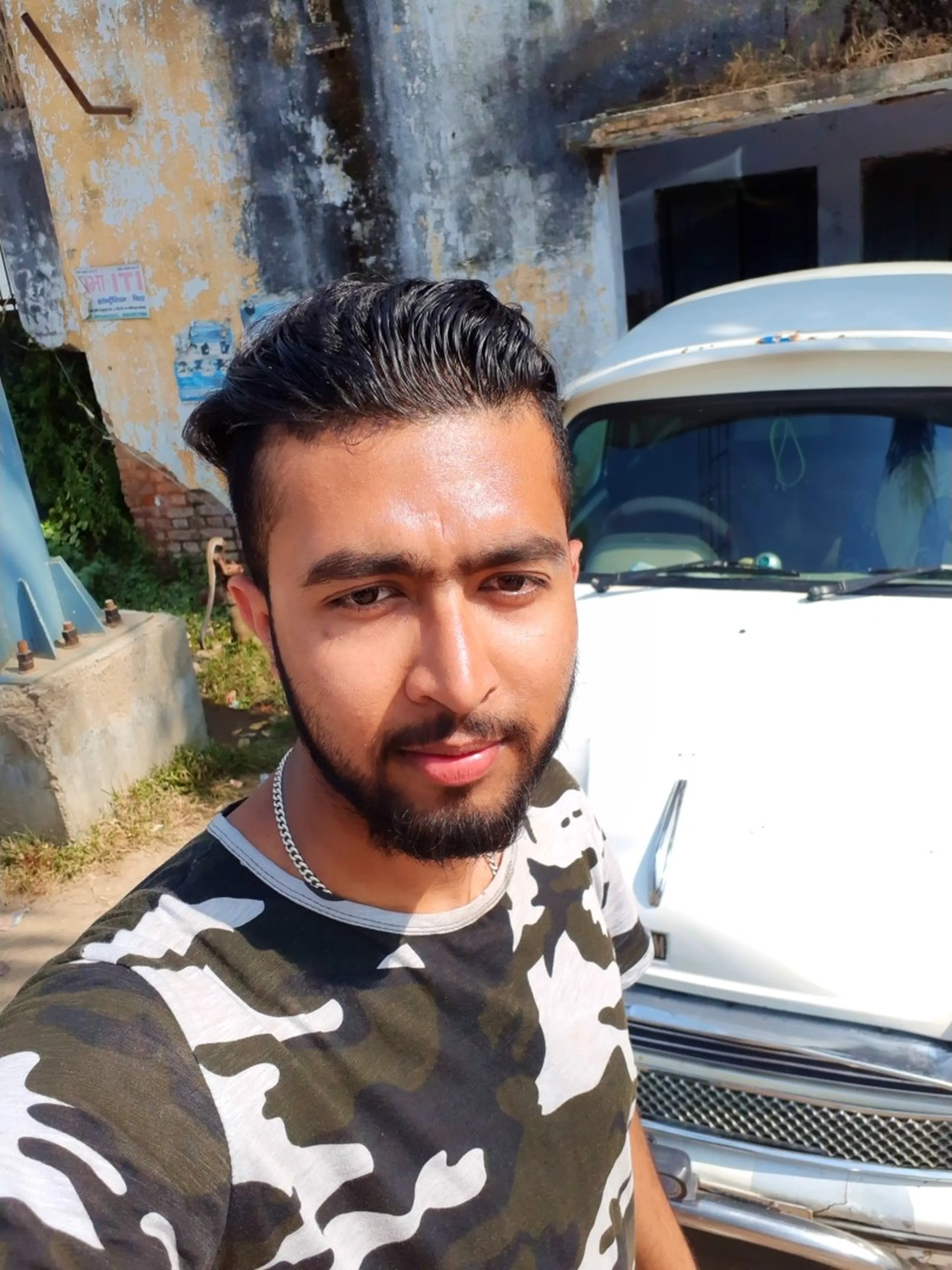 A close-up selfie portrait of a stylish young South Asian male trekker with a freshly styled slicked-back undercut hairstyle and a neatly trimmed short beard, wearing a grey and white camouflage-print short-sleeve t-shirt and a thin silver chain necklace, photographed in front of a weathered grey and yellow peeling paint concrete wall with a white classic Hindustan Ambassador car partially visible behind him to the right, captured during the Nepalgunj stopover between the remote Dolpa mountains and the return flight to Kathmandu, Nepalgunj Nepal post trek selfie, trekker fresh look Nepal lowlands, Hindustan Ambassador vintage car Nepal, Shey Phoksundo trek return Nepalgunj Nepal, same batch college trek Nepal, Nepal travel blog Nepalgunj