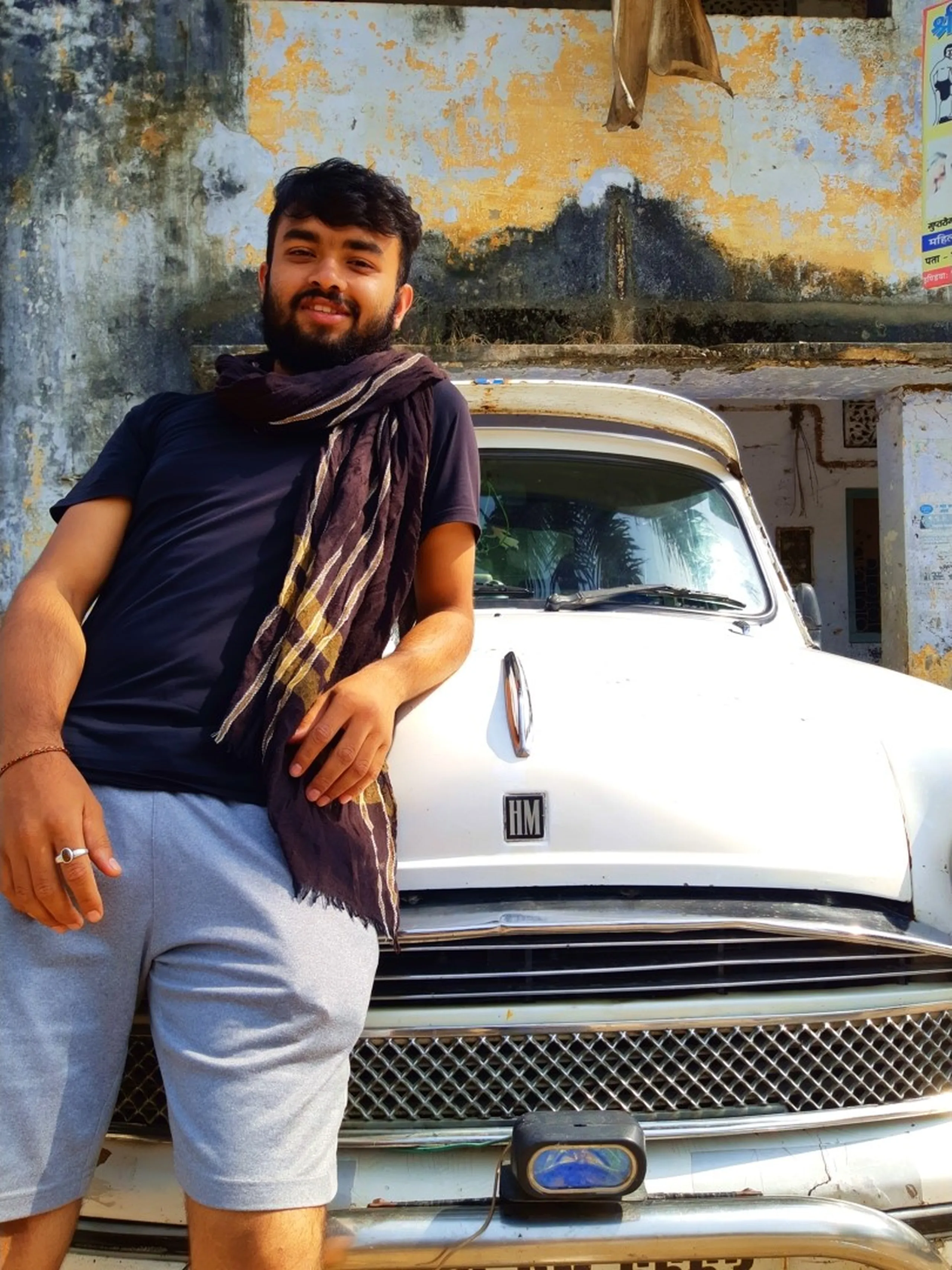 A relaxed and charming portrait of a bearded young South Asian male trekker leaning casually against the chrome grille and bonnet of a classic white Hindustan Ambassador HM car parked on a dusty Nepalgunj street, wearing a plain dark navy blue short-sleeve t-shirt, a dark brown and gold checked shawl or muffler draped around his neck, light grey shorts and a thin red string bracelet on his wrist, smiling warmly at the camera, a weathered peeling-paint concrete building wall with faded Devanagari text visible behind, the iconic HM badge of the vintage Ambassador clearly visible on the car bonnet, Hindustan Ambassador car Nepal Nepalgunj, trekker vintage car portrait Nepal, Shey Phoksundo trek return stopover Nepalgunj, Nepal classic car street photography, college batch mate trek Nepal lowlands, Nepal adventure travel return journey