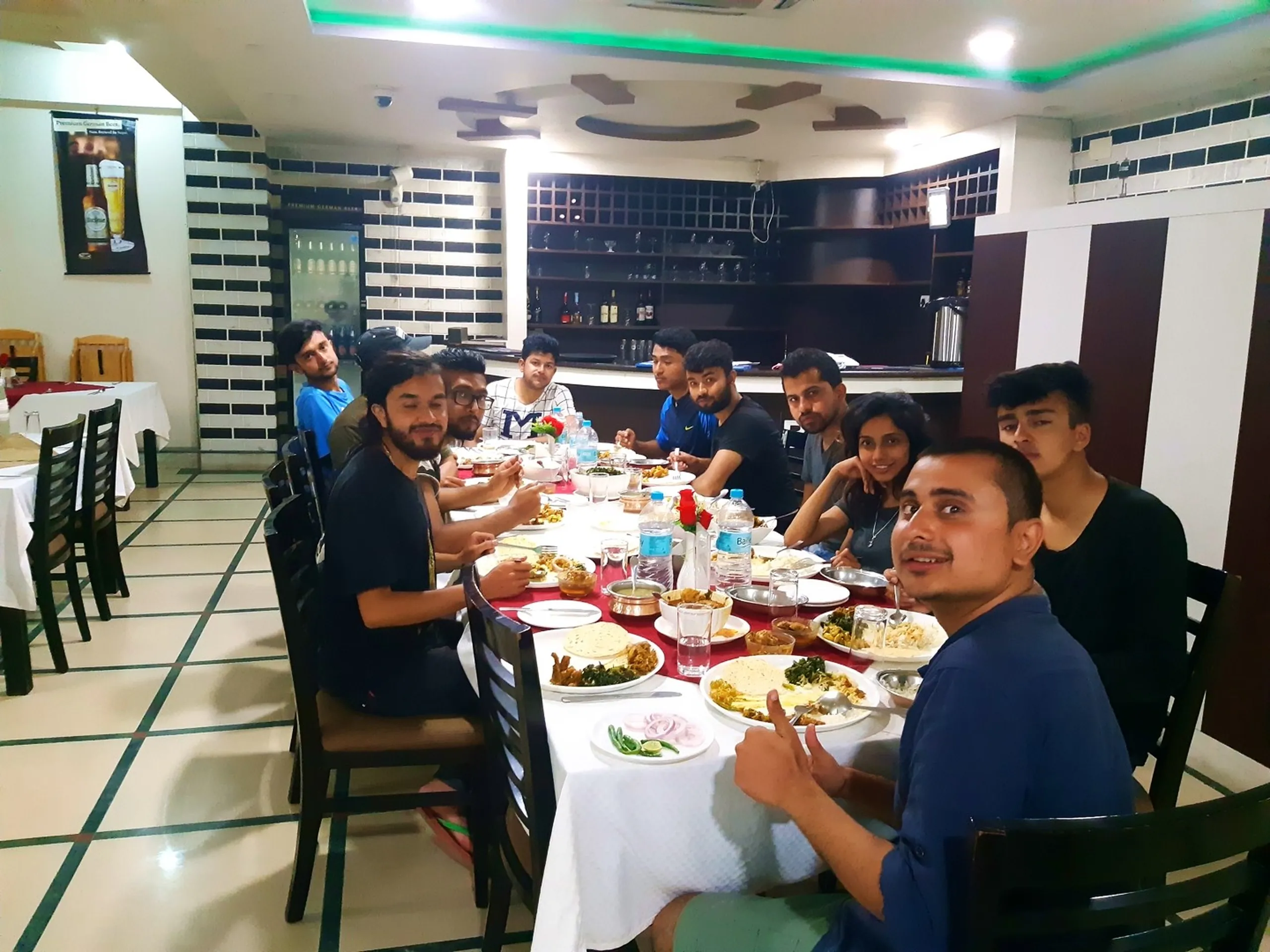 A wide indoor group dinner photograph of the entire Shey Phoksundo trekking group seated around a long rectangular restaurant table covered in a white tablecloth at a well-lit modern Nepalgunj restaurant, celebrating the completion of their epic Dolpa trek - approximately eleven people including men and women in casual post-trek clothing (blue t-shirts, black jackets, a grey graphic tee) seated in dark wooden chairs, the long table filled with multiple steel bowls of curry, plates of bread, bottled water, glasses and a small red rose vase centerpiece, a glass-front bar unit with trophy glasses and liquor bottles visible along the back wall with a beer brand poster, green LED accent lighting along the ceiling cornice, the atmosphere one of joyful exhausted celebration, Shey Phoksundo trek completion dinner Nepalgunj, trekking group restaurant celebration Nepal, college friends post trek dinner Nepal, Dolpa trek group Nepal celebration, Nepalgunj restaurant trekkers Nepal, batch mates group dinner Nepal adventure