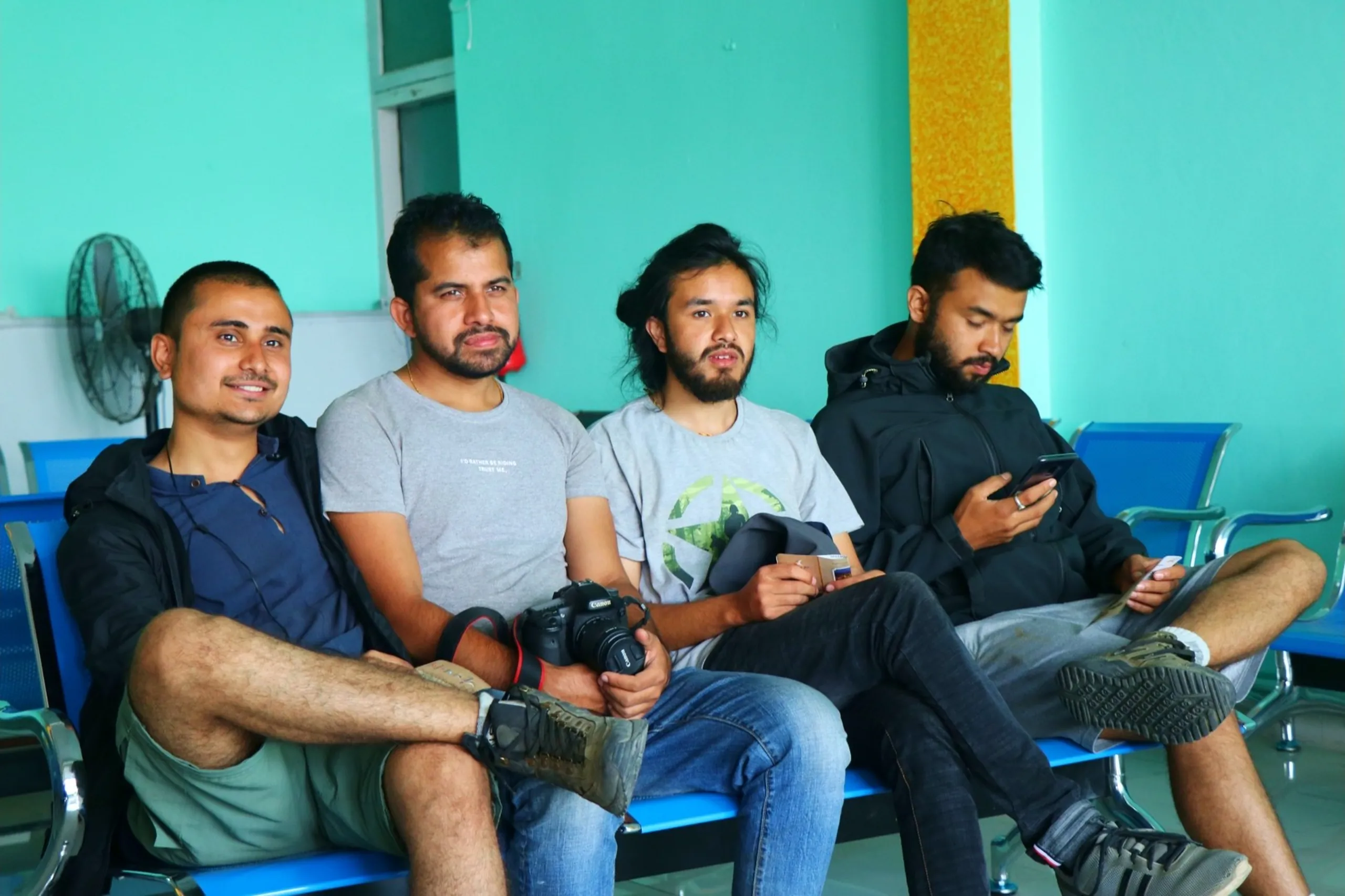 Four young South Asian male trekkers seated in a row on blue airport waiting chairs in a bright turquoise-walled departure lounge, waiting to board their flight from Nepalgunj to Kathmandu - from left to right: one with a buzz cut and visible ear piercing in a black jacket smiling slightly, the second in a light grey "I'd Rather Be Riding - Trust Me" graphic t-shirt with a short beard and gold chain necklace holding a Canon DSLR camera on his lap, the third with long hair tied in a bun and a beard in a grey graphic t-shirt holding a small item and looking directly at the camera, and the fourth in a dark black hoodie jacket scrolling on a smartphone with legs crossed, worn hiking shoes visible, a pedestal fan and a yellow textured wall panel in the background, Nepalgunj airport departure lounge Nepal, trekkers waiting board flight Nepal, Canon DSLR photographer trekker Nepal, batch mates college friends Nepalgunj airport, post Dolpa trek flight Kathmandu Nepal, Nepal domestic flight Nepalgunj Kathmandu