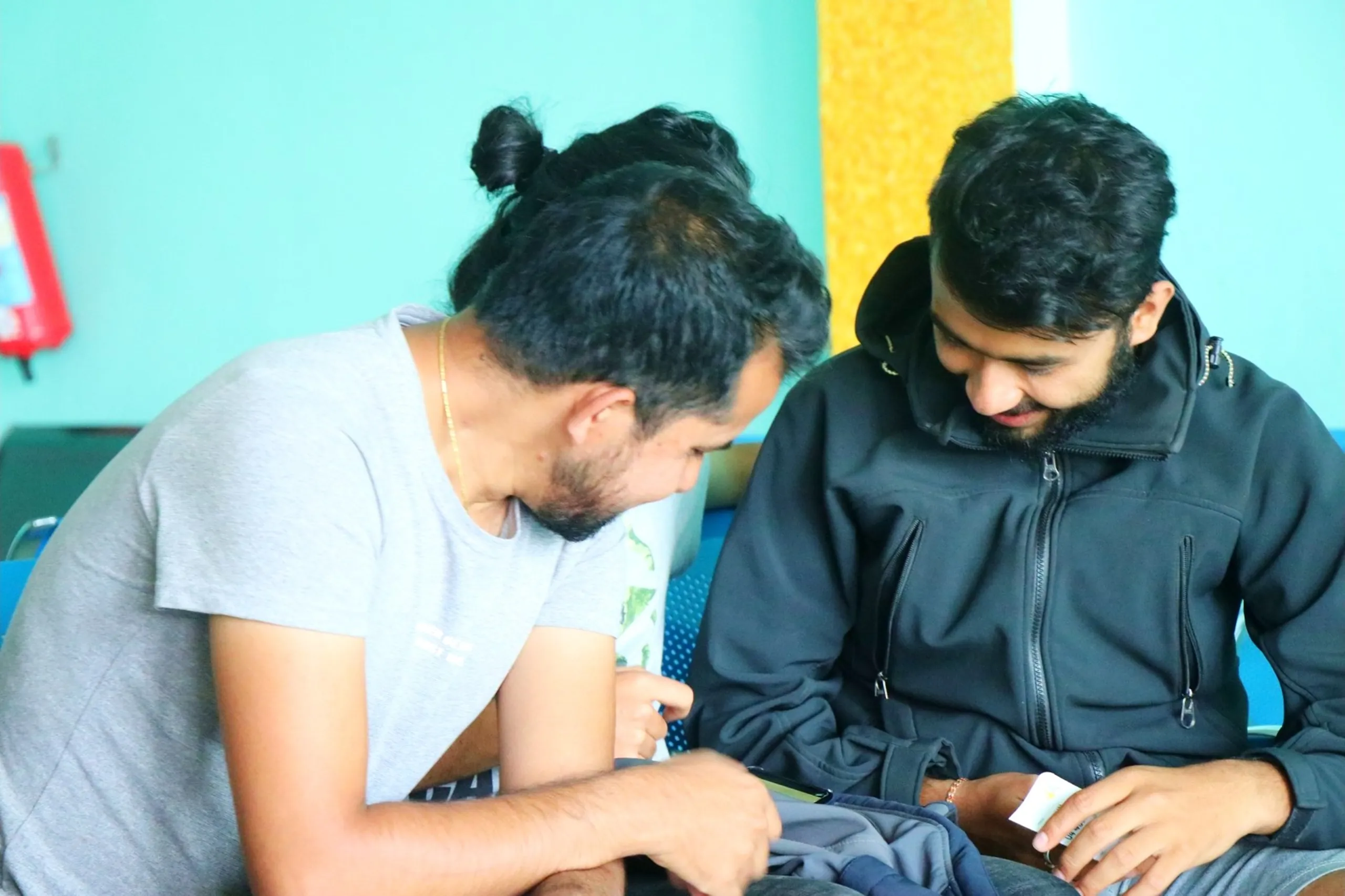 A candid and intimate close-up photograph of two young South Asian male trekkers leaning closely together in the turquoise-walled airport waiting lounge, one with a man-bun hairstyle and beard in a light grey graphic t-shirt leaning in intently to look at something being shown by the other - a bearded trekker in a dark black zip-up hooded jacket who appears to be showing a small sticker, badge or card object with a subtle smirk, both their expressions radiating shared curiosity and amusement, a red fire extinguisher bracket partially visible on the wall to the left, a yellow textured column behind, Nepalgunj airport waiting lounge Nepal candid, batch mates sharing moment Nepal airport, trekking friends bonding Nepal Nepalgunj, post trek Nepal domestic airport, Shey Phoksundo trek friends Kathmandu flight, authentic Nepal travel photography candid