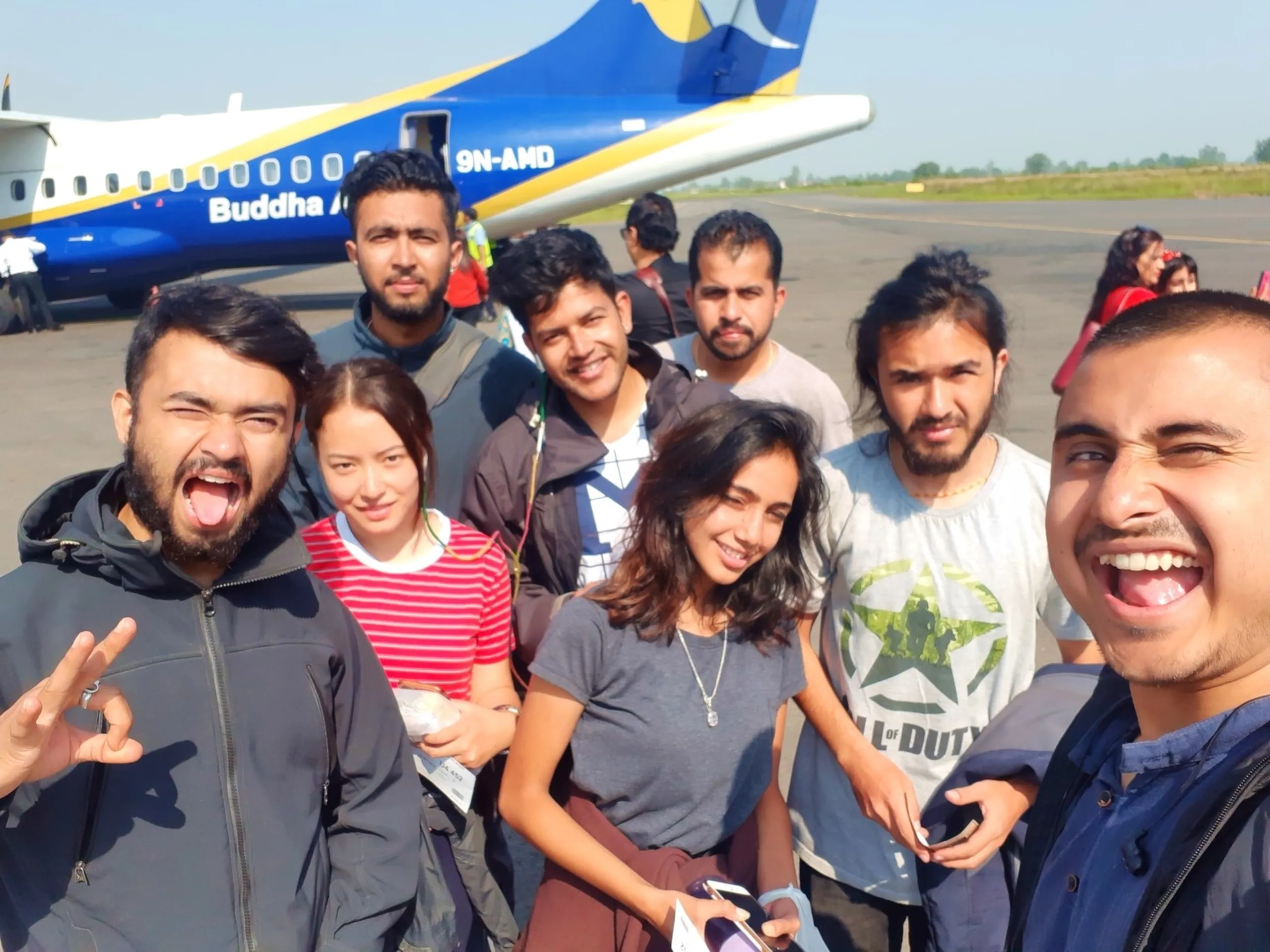 An exuberant and chaotic group selfie of the Shey Phoksundo trekking family moments before boarding their Buddha Air flight at Nepalgunj Airport - eight people crowding into the frame on the sun-drenched tarmac with the distinctive blue and yellow Buddha Air ATR turboprop aircraft registration 9N-AMD looming directly behind them - two trekkers in the front row sticking their tongues out wildly with one flashing a peace sign, a laughing woman in a red and white striped t-shirt, another in a grey t-shirt holding boarding passes and smiling, a bearded trekker in a grey Call of Duty graphic tee, one with a man-bun and a short gold necklace, and two more in the back row - the entire image radiating the raw joy and bittersweet energy of a journey coming to its end, Buddha Air Nepal Nepalgunj airport group selfie, Shey Phoksundo trek final selfie Nepal, trekking group last photo Nepal airport, Buddha Air 9N-AMD Nepal domestic flight, college batch mates trek farewell Nepal, Nepal adventure trek completion group photo
