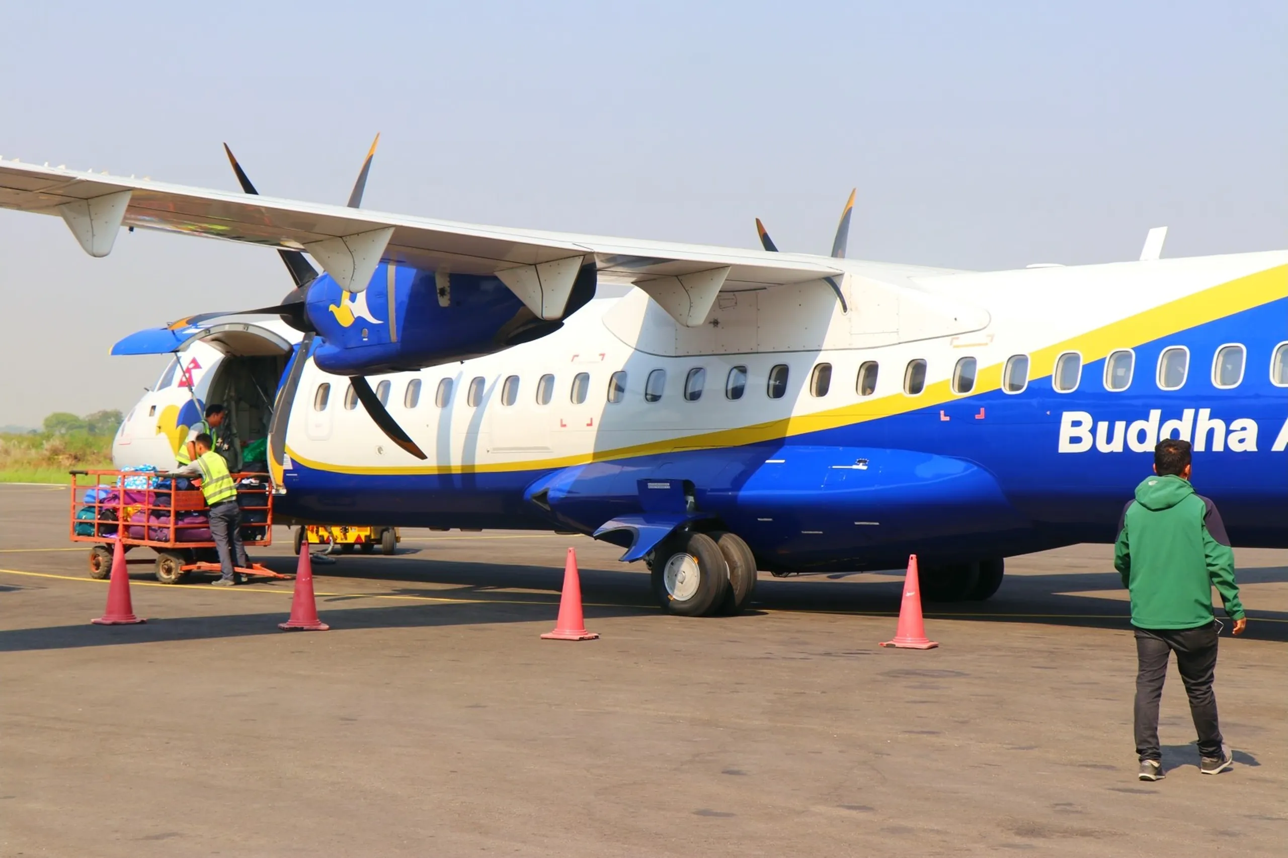 A clean and professional tarmac-level photograph of a Buddha Air ATR-72 twin-turboprop passenger aircraft parked on the flat open tarmac of Nepalgunj Airport under a hazy pale grey sky, showing the full left side of the aircraft in its bold blue, white and yellow corporate livery with "Buddha A..." text visible on the fuselage, the large blue engine nacelle and spinning propeller in view, two ground crew workers in yellow safety vests loading colorful luggage onto an orange baggage trolley cart through the forward cargo hold, orange traffic safety cones arranged below the wings, and one trekker in a green jacket walking toward the aircraft steps in the right background, Buddha Air Nepal domestic flight, Buddha Air ATR72 Nepalgunj airport, Nepal domestic aviation, Nepalgunj Kathmandu flight Nepal, domestic airline Nepal Shey Phoksundo trek return, Nepal airport tarmac photography